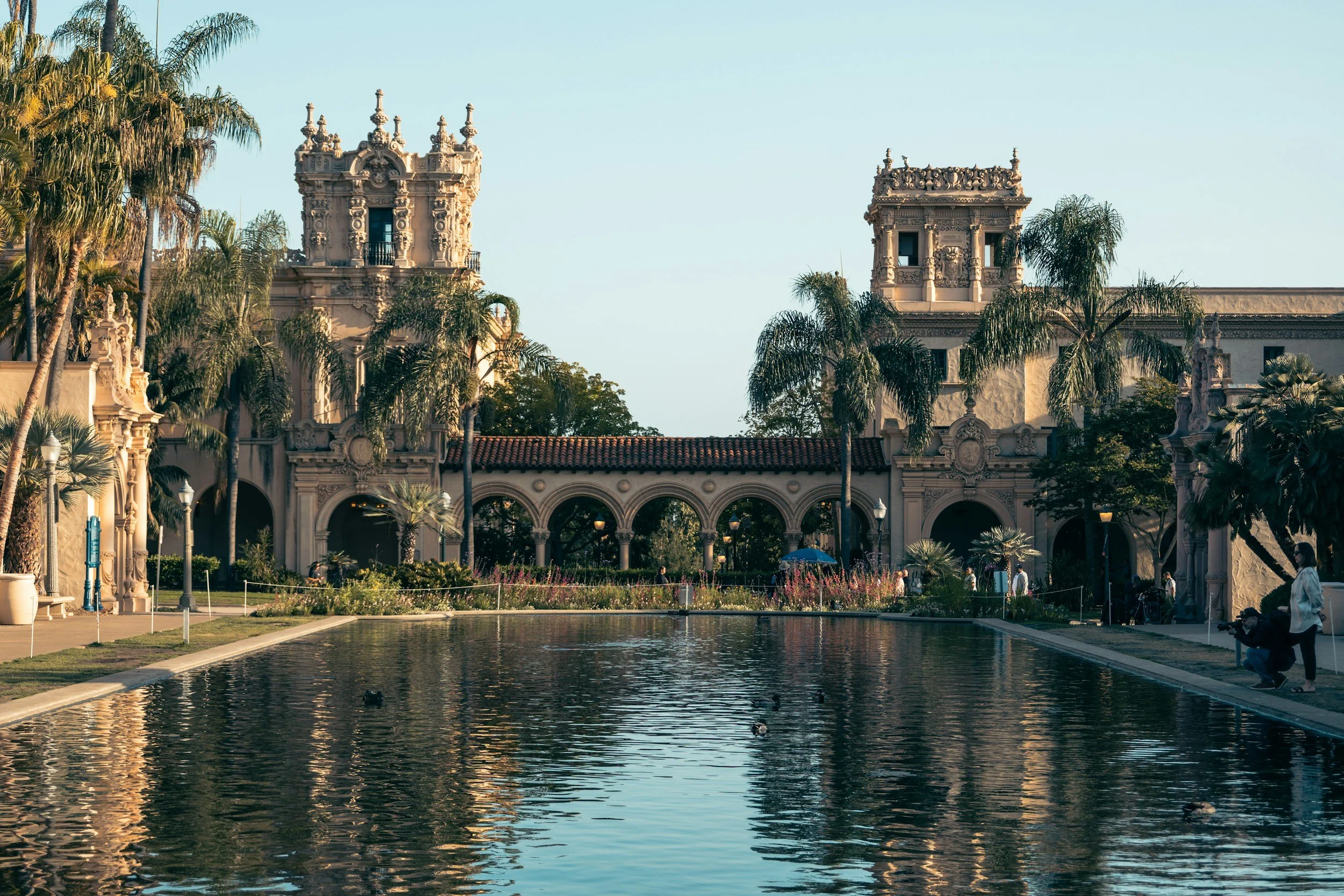 The House of Hospitality in Balboa Park, San Diego can be seen reflected in a pond. The Spanish revival building is ornate, with two towers connected by a walkway. It is surrounded by palm trees