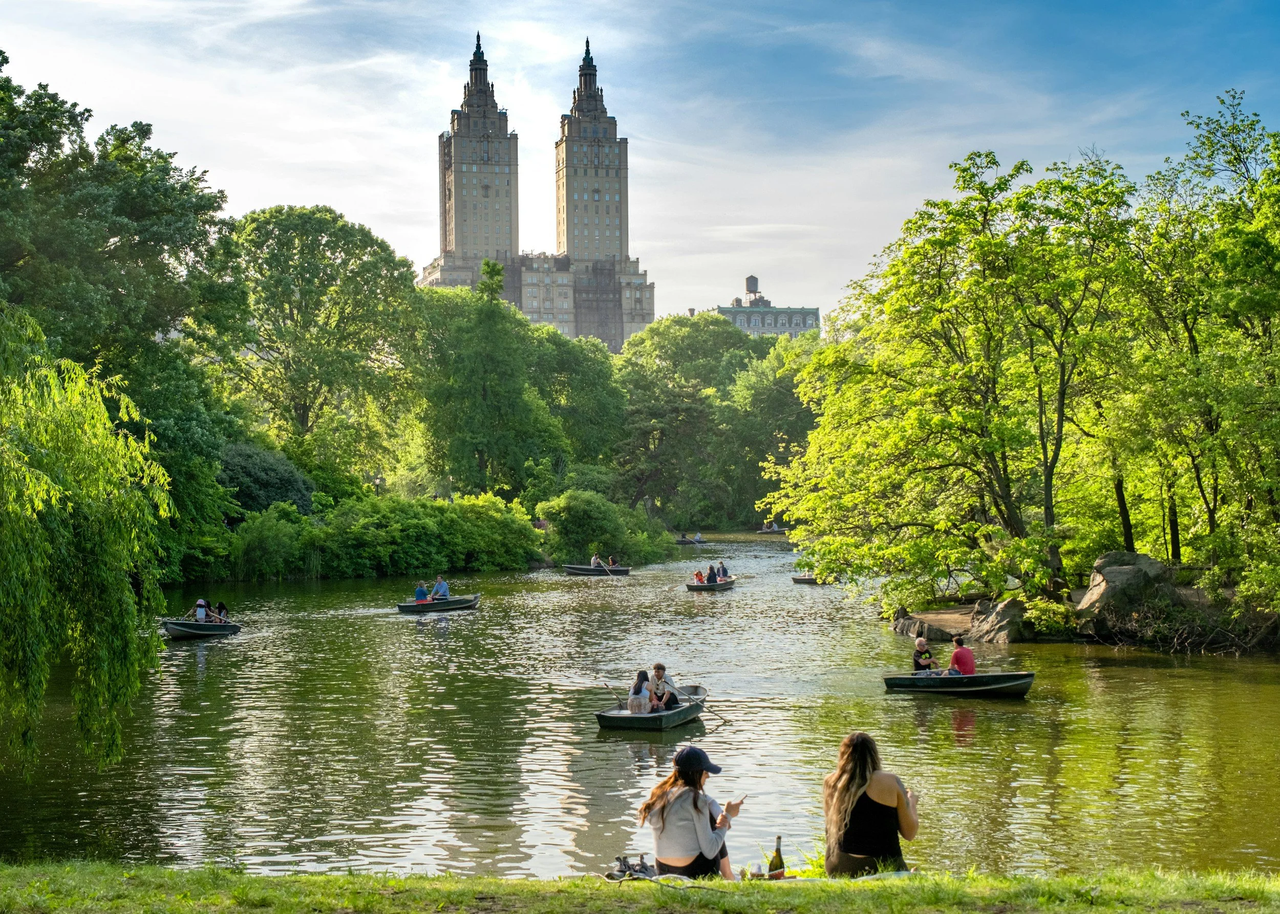 People sit and watch rowing boats on a lake in Central Park, New York. The lake is surrounded by green trees, with skyscrapers in the distance