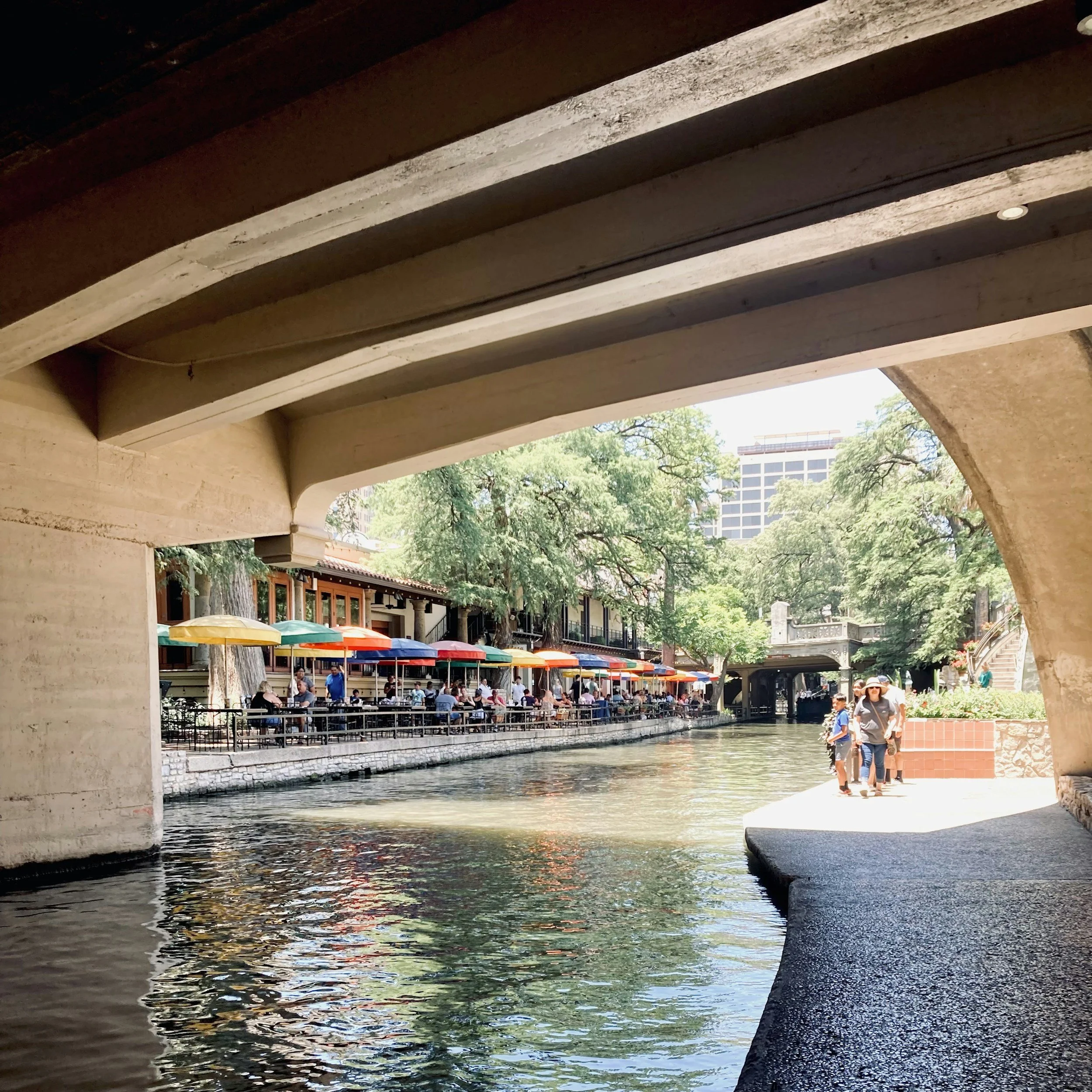 People walk along the edge of the San Antonio River Walk, which is lined on the opposite bank by cafes and restaurants