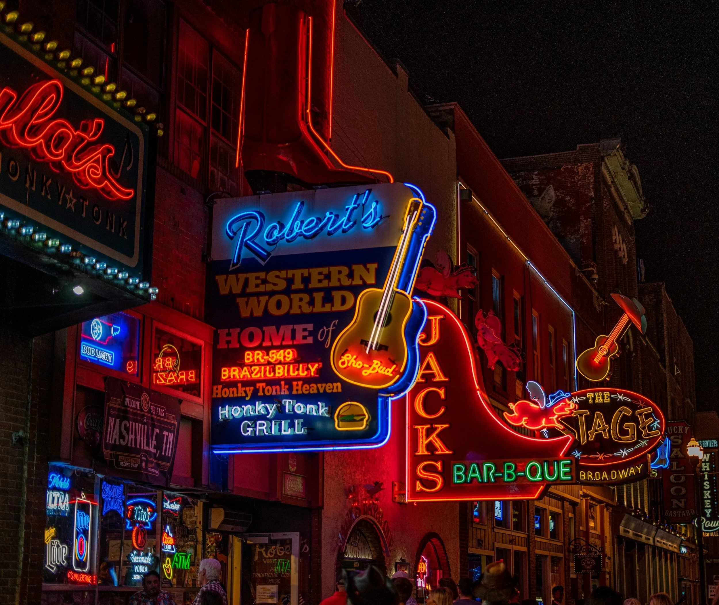 The bright neon lights of Downtown Nashville shine at night. The focus is on a huge guitar hanging outside Robert's Honky Tonk Grill