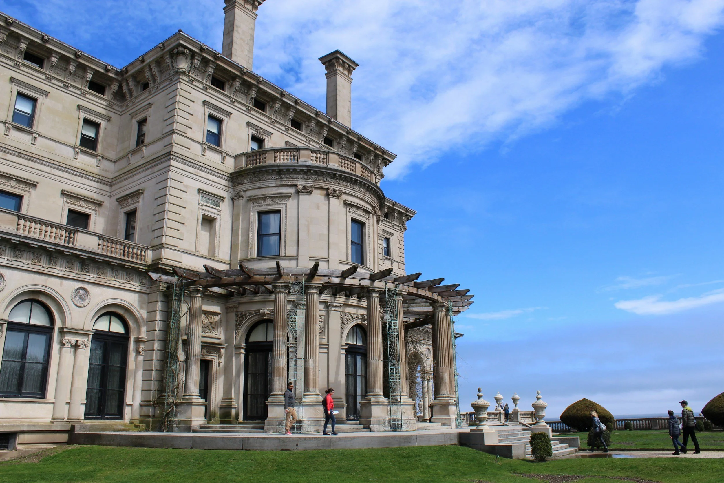 People walk around the porch of the Breakers under a blue sky. It towers over them. Drives & Detours Newport Cliff Walk tips