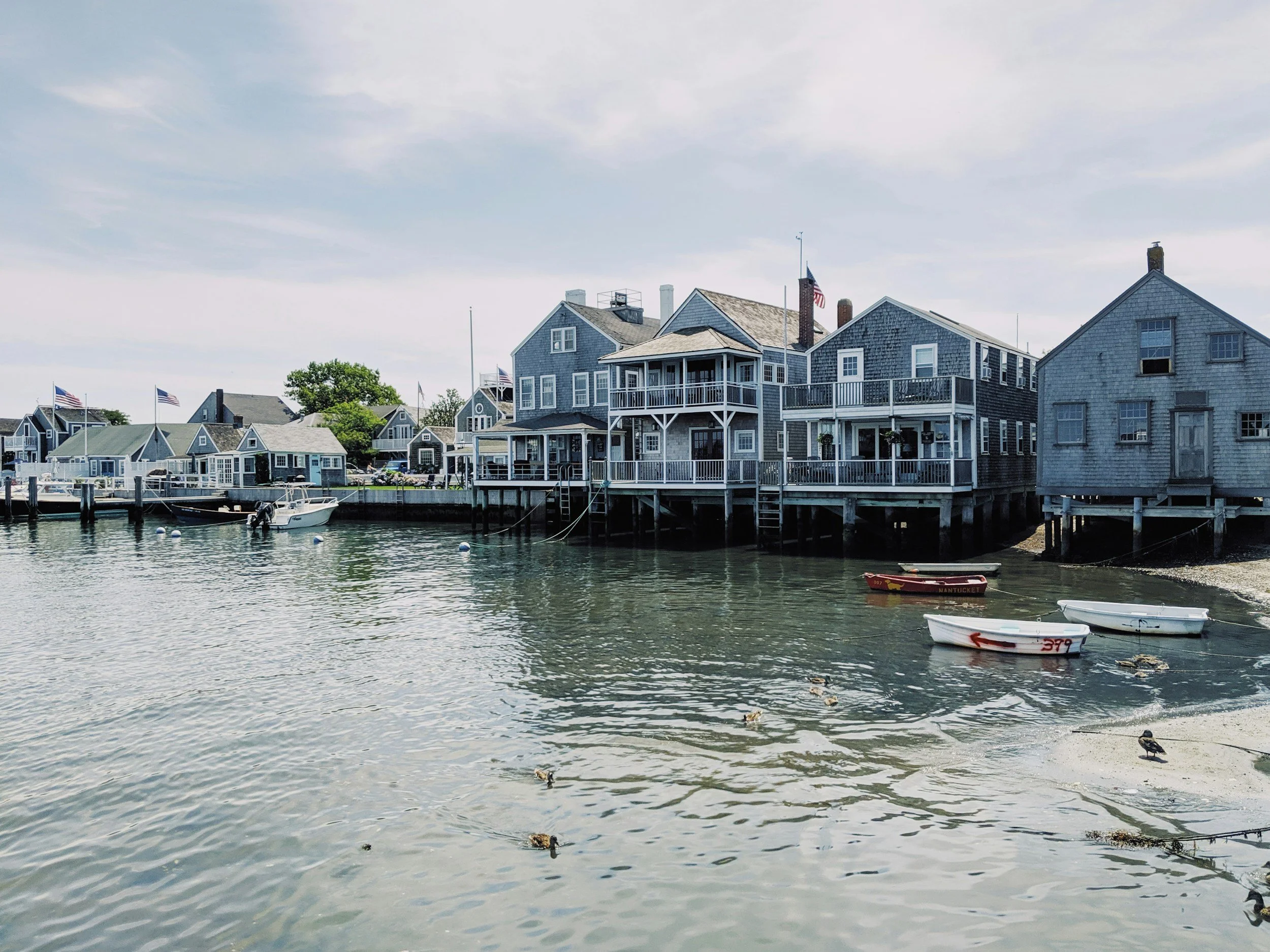 Wooden houses line a beach with small boats floating in still water. Drives & Detours Nantucket Tour Bundle