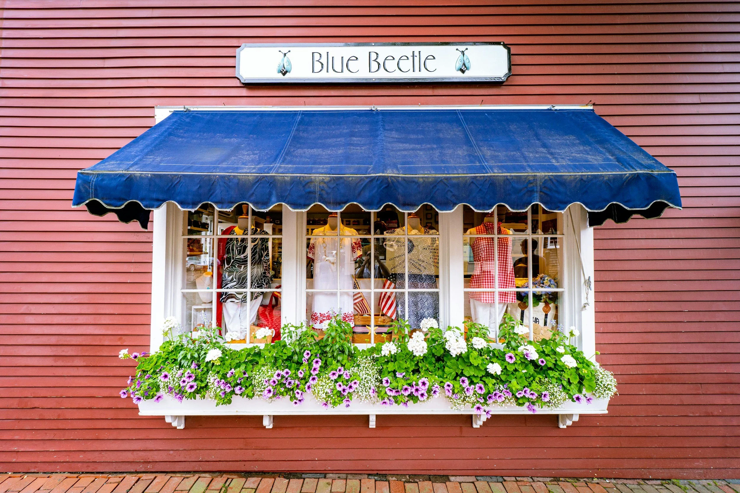 Flower baskets line a window with a blue awning. The window is full of clothes and mannequins, and in a red wooden wall. Drives & Detours Nantucket Tour Bundle