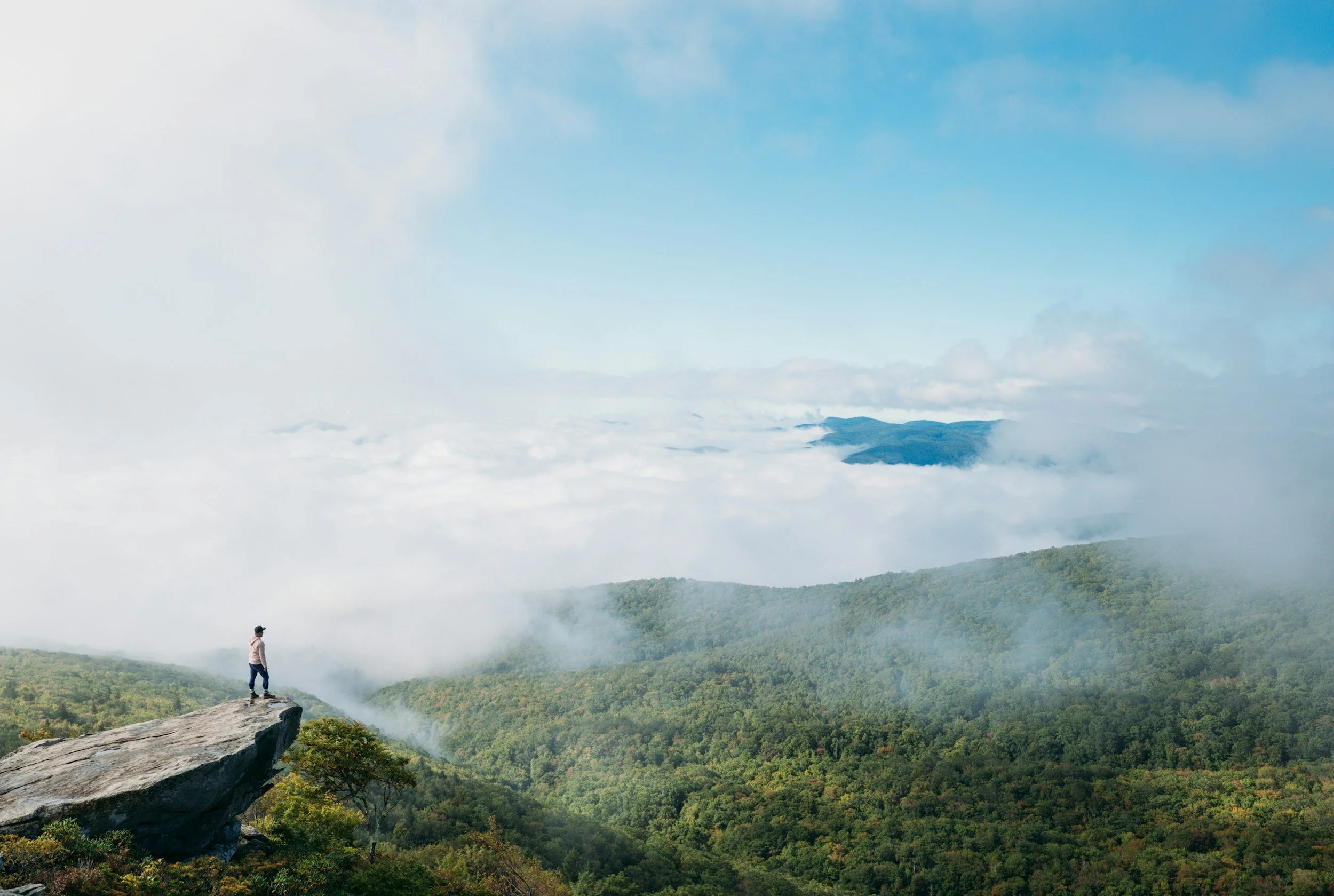 A man stands on a large rock overlooking a green valley full of clouds in Tennessee. Drives & Detours Great Smoky Mountains first time