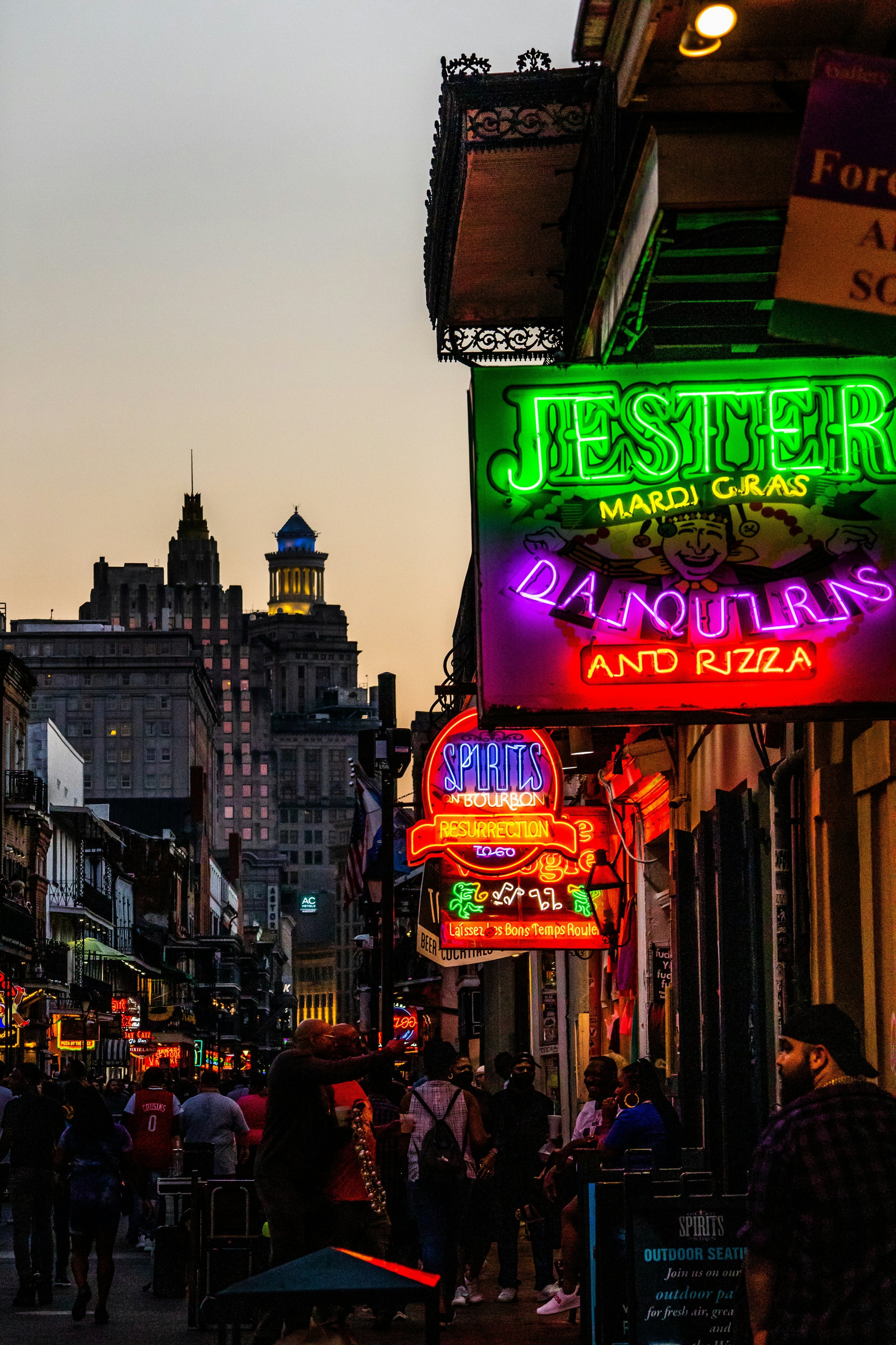 Bourbon Street in New Orleans is lit up by neon signs offering drinks at dusk. The street is full of people