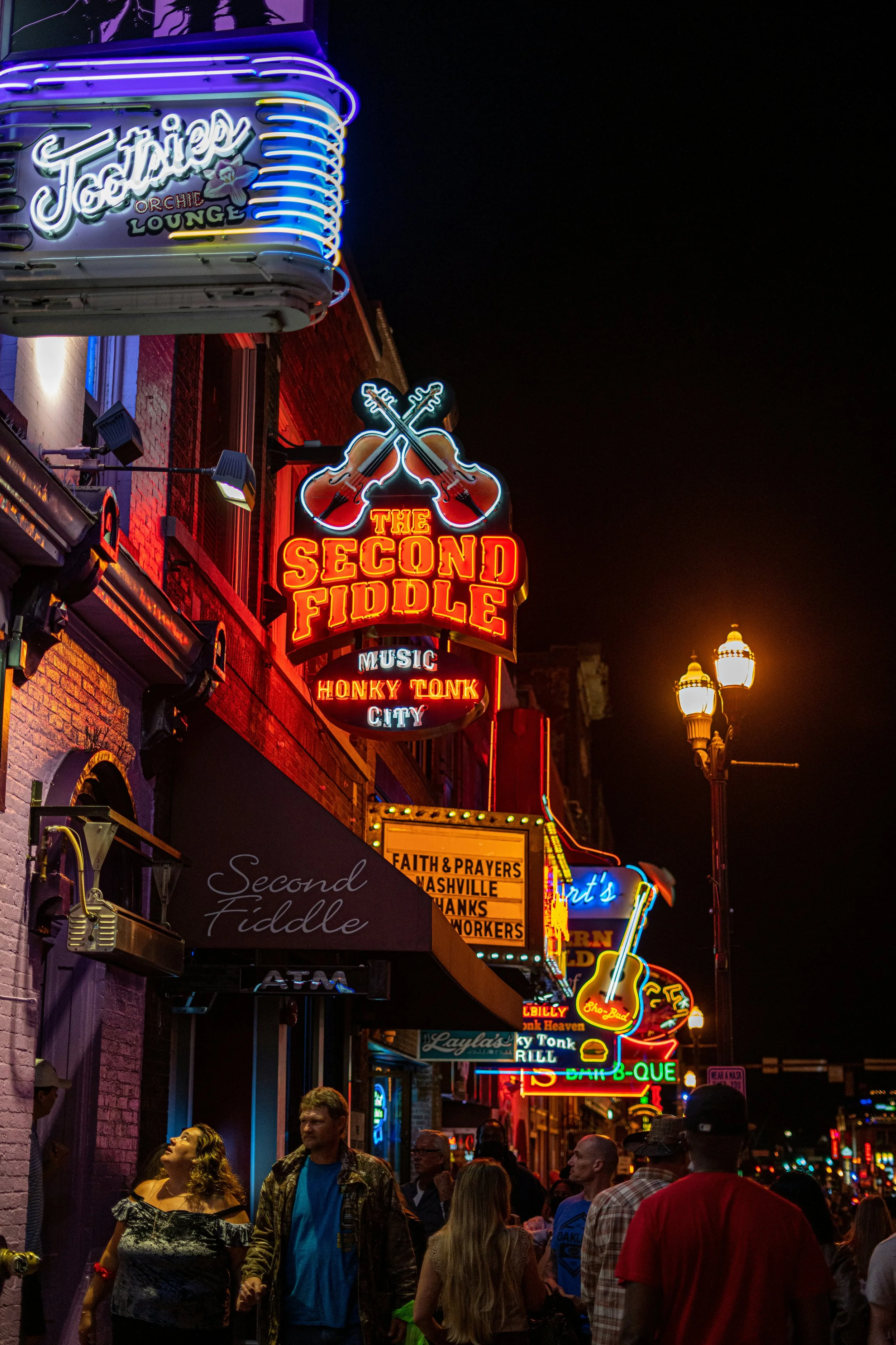 Broadway in Nashville, Tennessee, after dar. The neon signs are lit up brightly over the heads of the people walking underneath. Drives & Detours Nashville self-guided tour