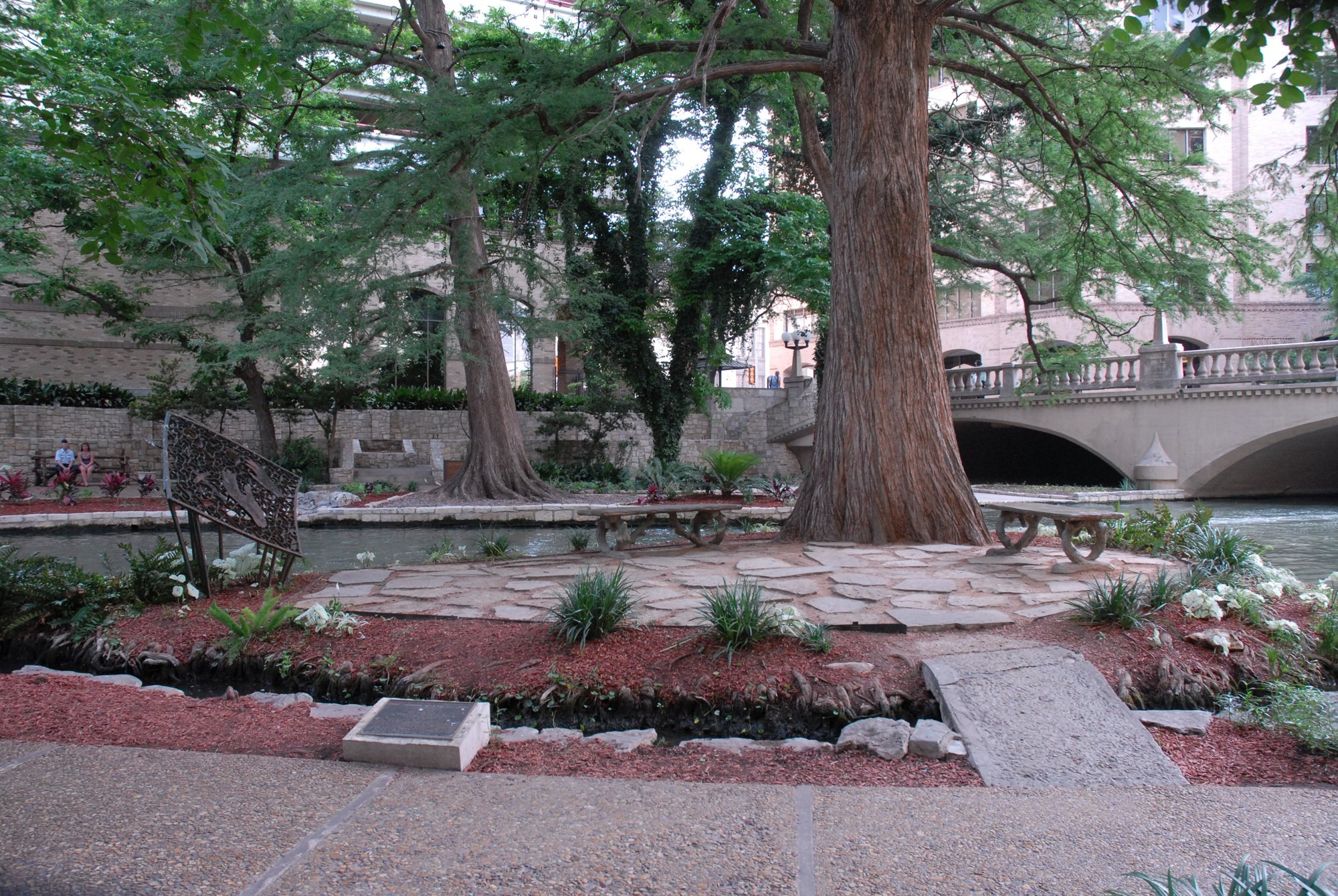 A small island, paved with benches and a tree, in a river. A bridge is in the background. Drives & Detours San Antonio River Walk hidden gems