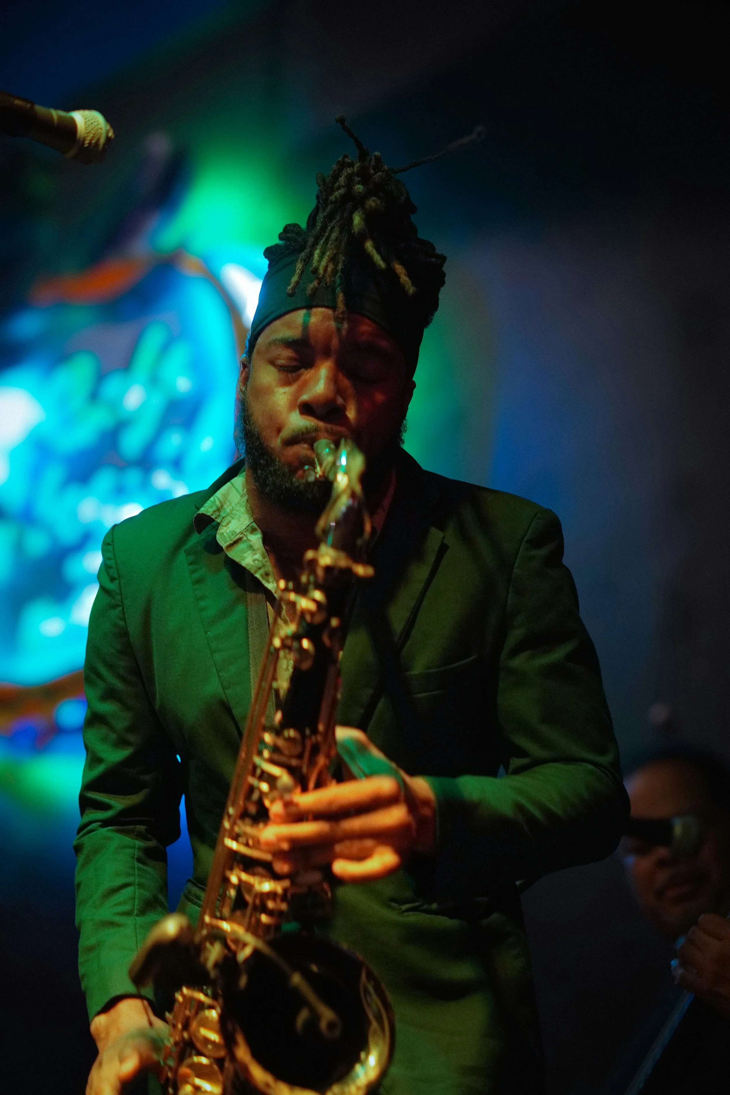 A man plays a saxophone in a jazz club. He's wearing a dark suit and his hair is tied up in dreadlocks. His eyes are closed as he he plays. The club is dark, and lit with green and blue light