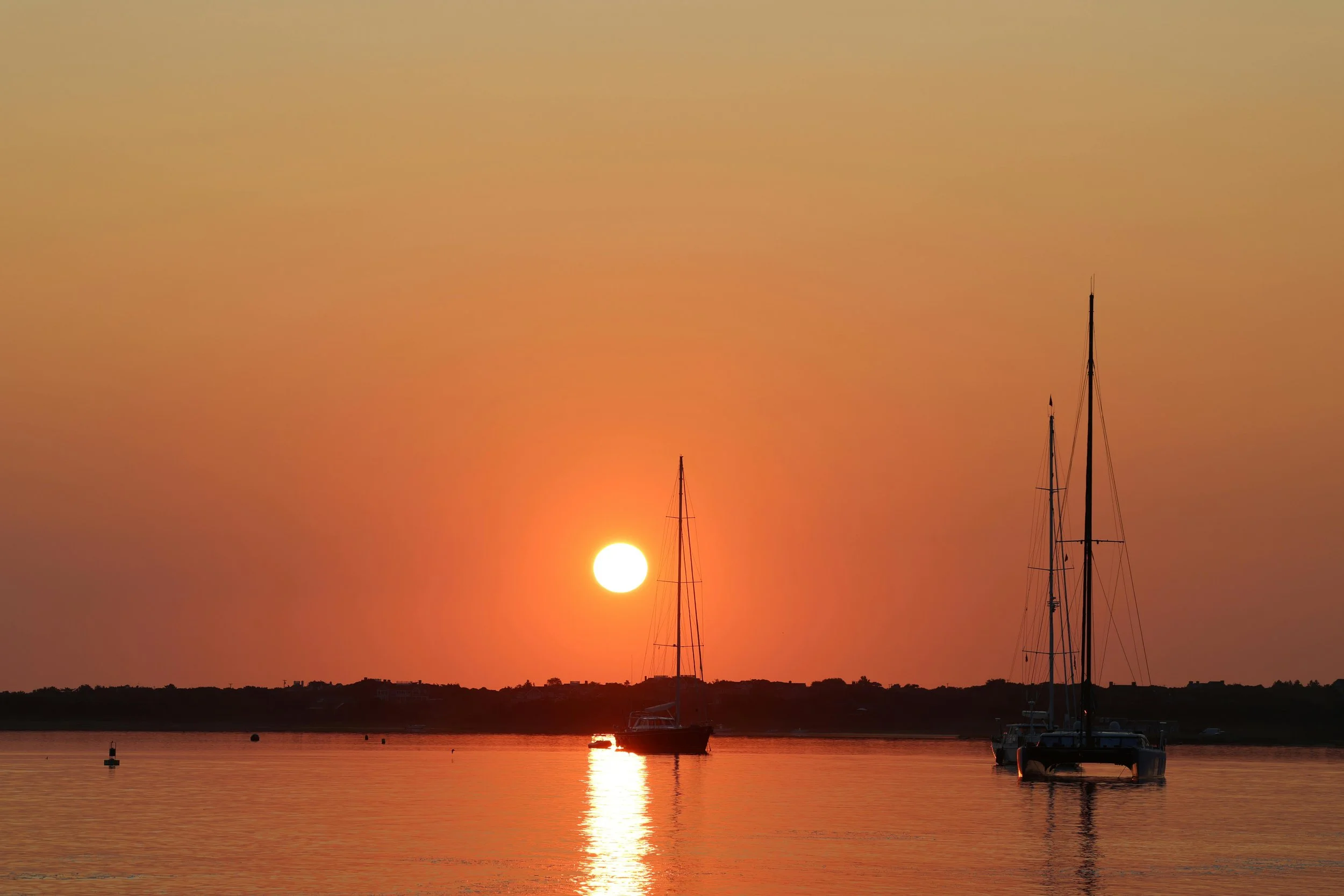 Sunrise from Brandt Point Lighthouse. A yacht is silhouetted by the low, orange sun. The boat is breaking the long reflection of the sun in the water. The sky is a deep orange