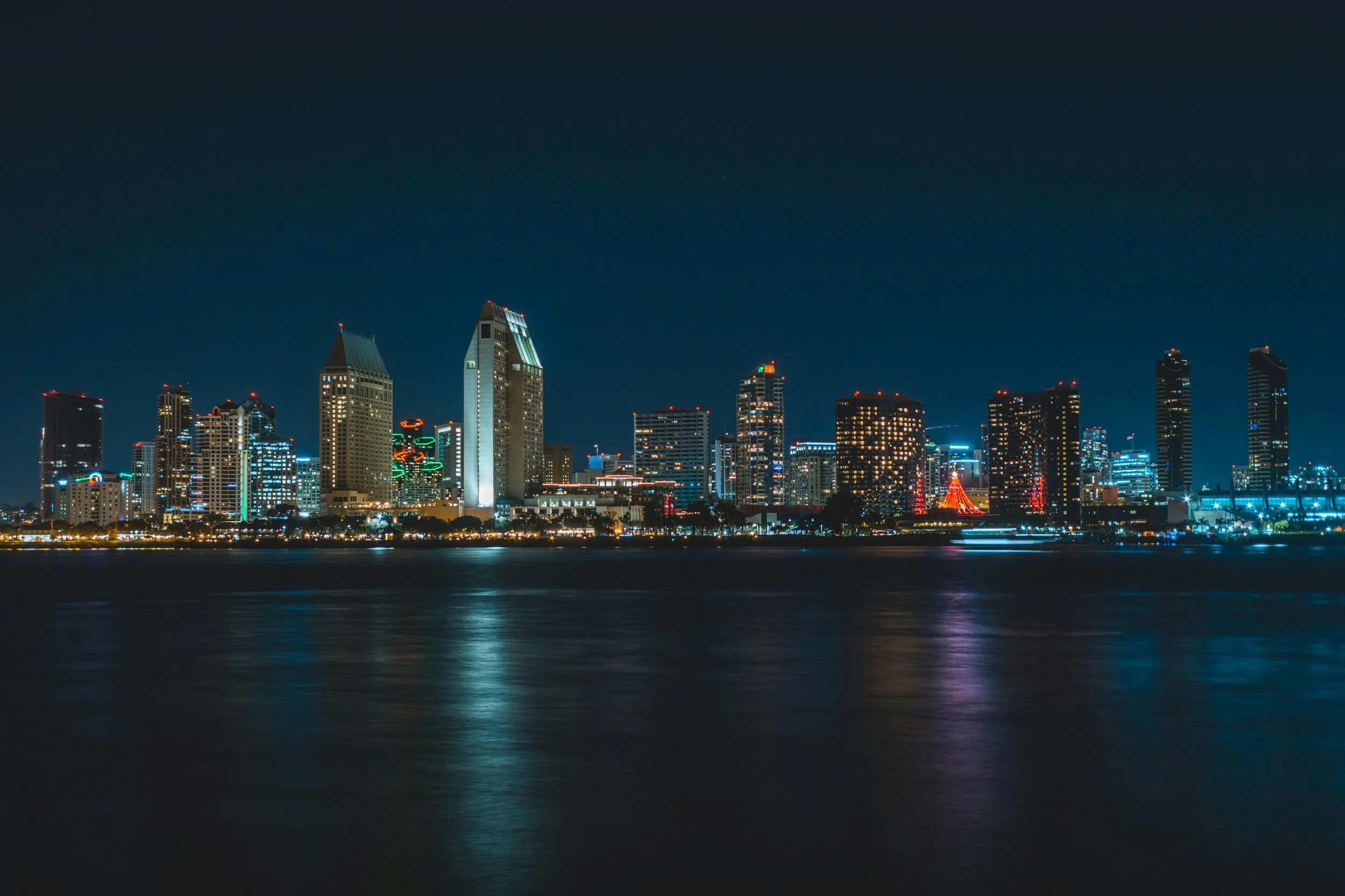 The San Diego skyline lights up at night, and is reflected in the bay in front. Drives & Drtours San Diego Embarcadero foodie guide
