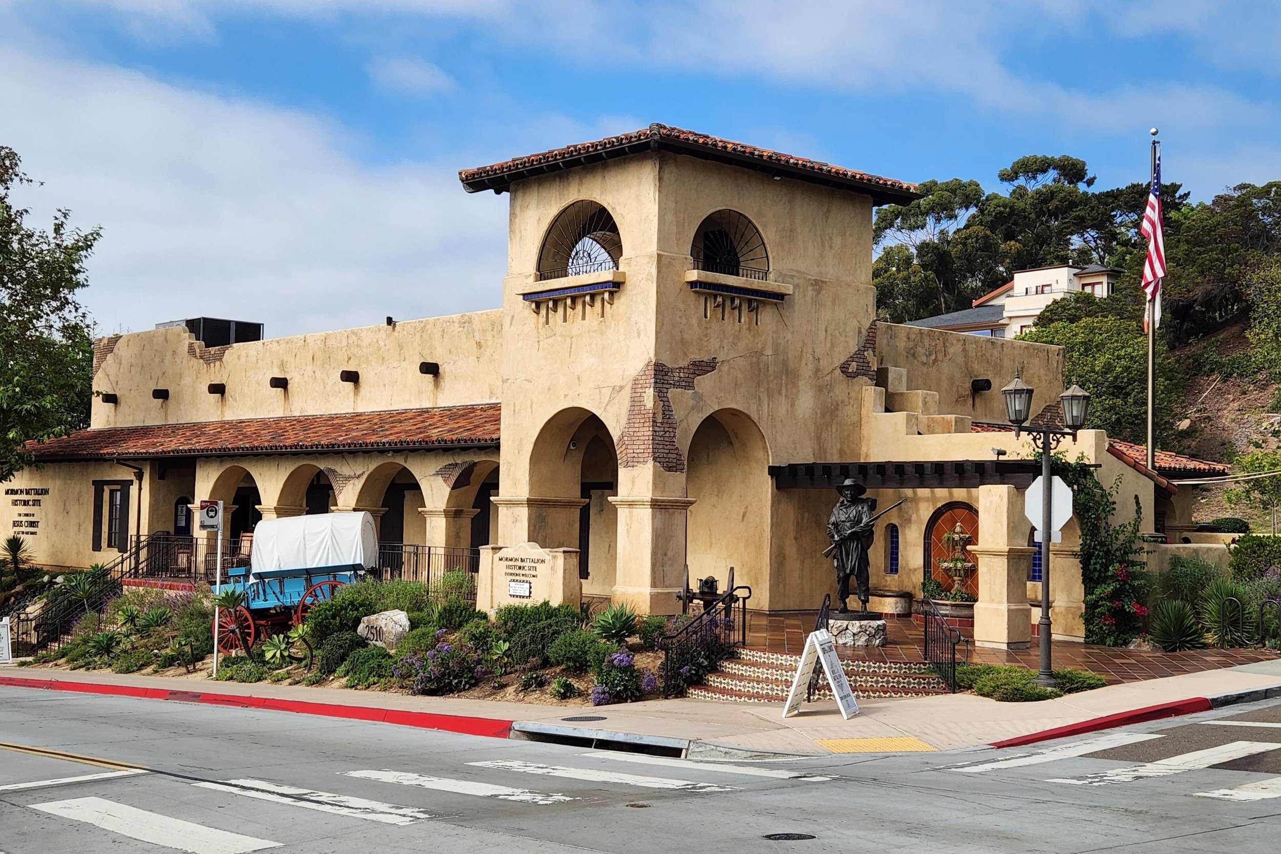 An adobe building that marks the site of the Mormon Battalion Historic Site. A statue outside shows a man carrying a rifle, and there is a blue wagon with red wheels