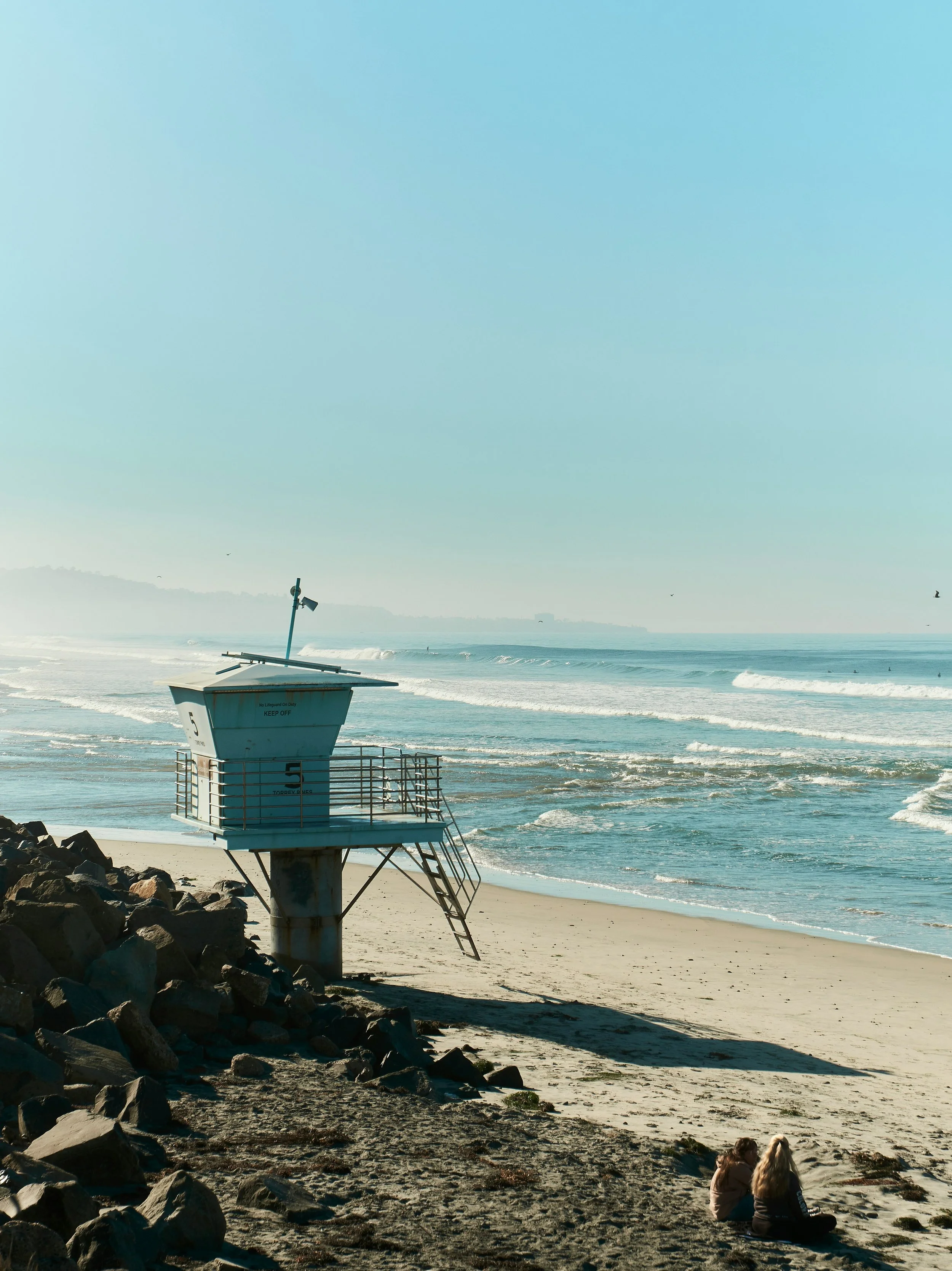 Two people sit beside a lifeguard hut at Torrey Pines Beach. The sand is smooth and large waves break in the distance