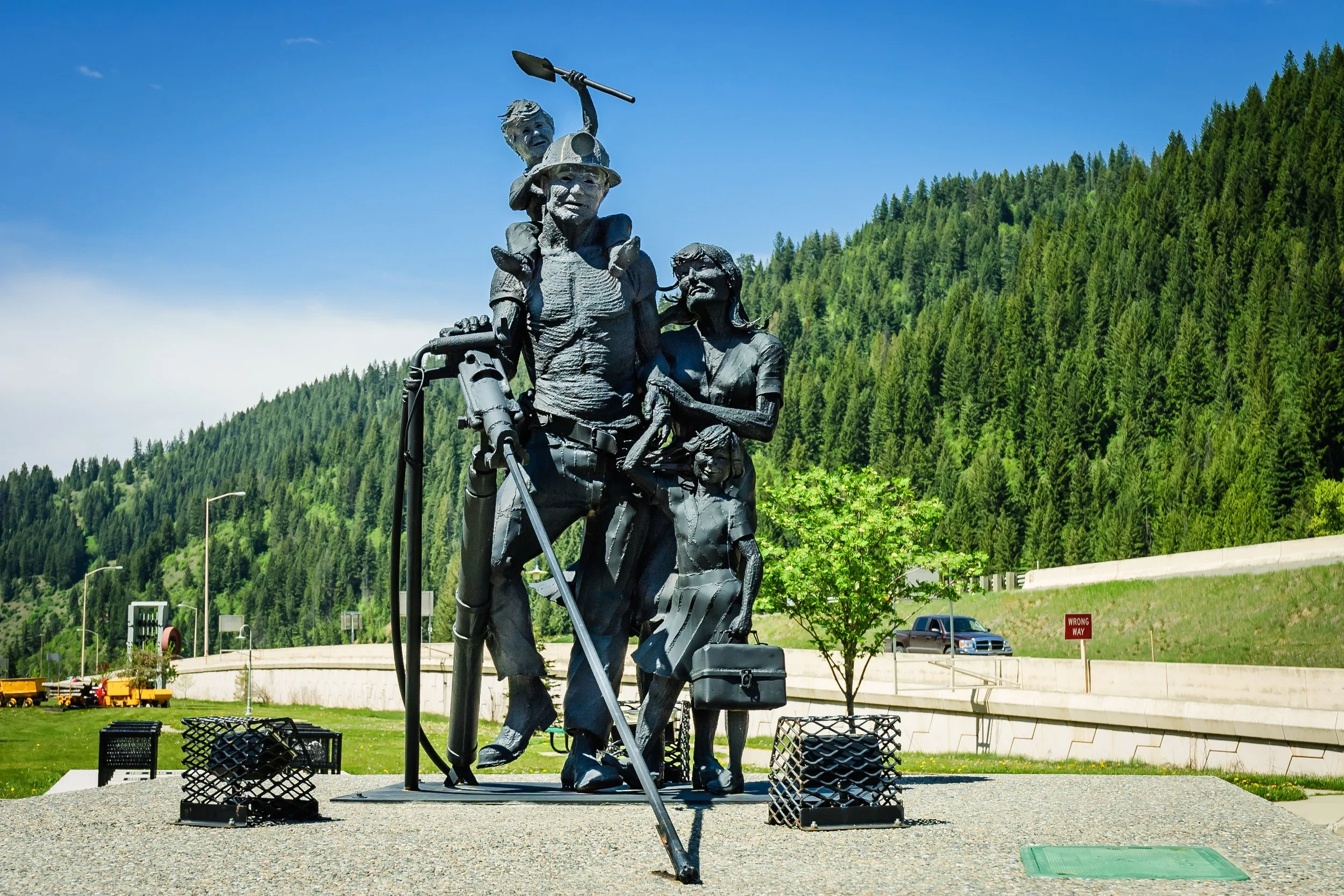 The Chamber Of Commerce Miner Family Statue in Idaho can be seen against a background of green fir trees. Drives & Detours Wallace tour