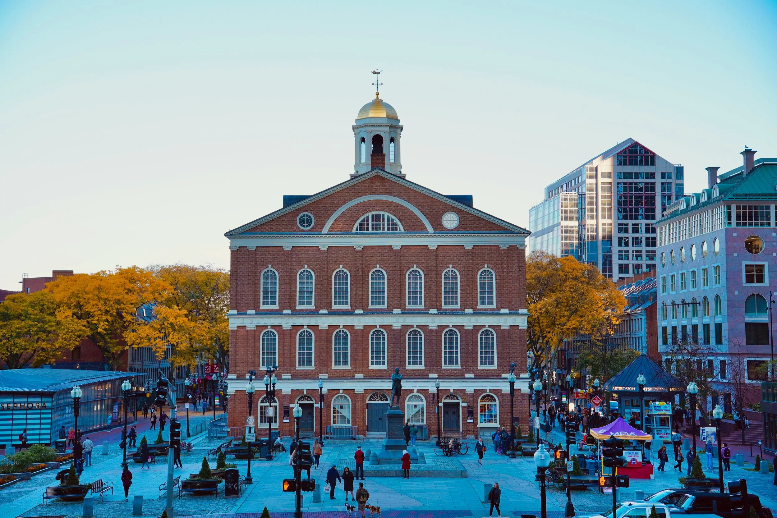Faneuil Hall as the sun sets over Boston. The large brick marketplace has a small golden dome on its roof. People mill about in the square in front