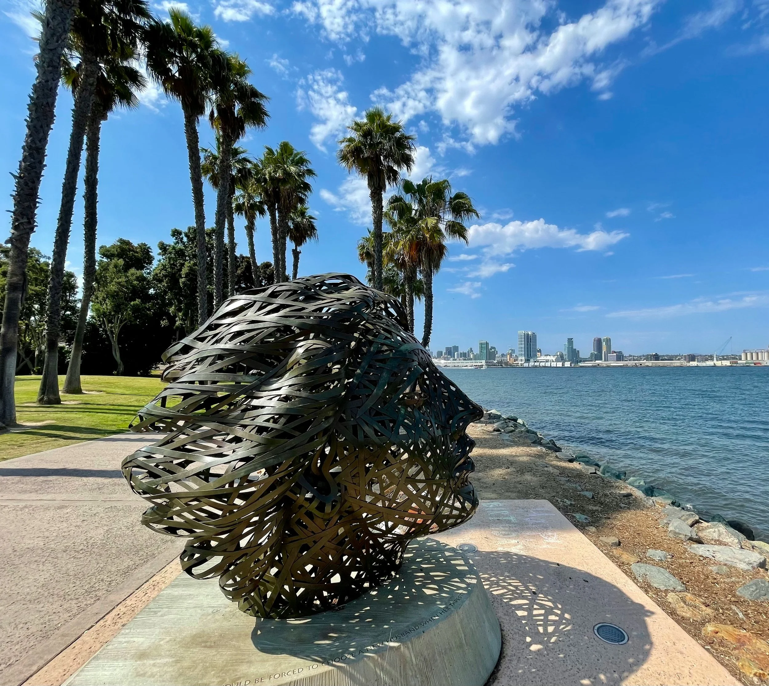 A sculpture of a head on the seafront of Coronado Island. It is surrounded by trees and the San Diego skyline can be seen in the background