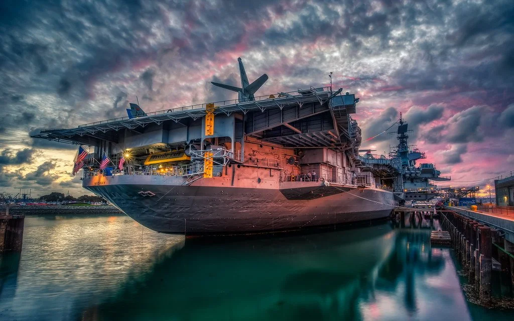 The USS Midway Museum in San Diego is lit up red at dusk, and surrounded by angry clouds