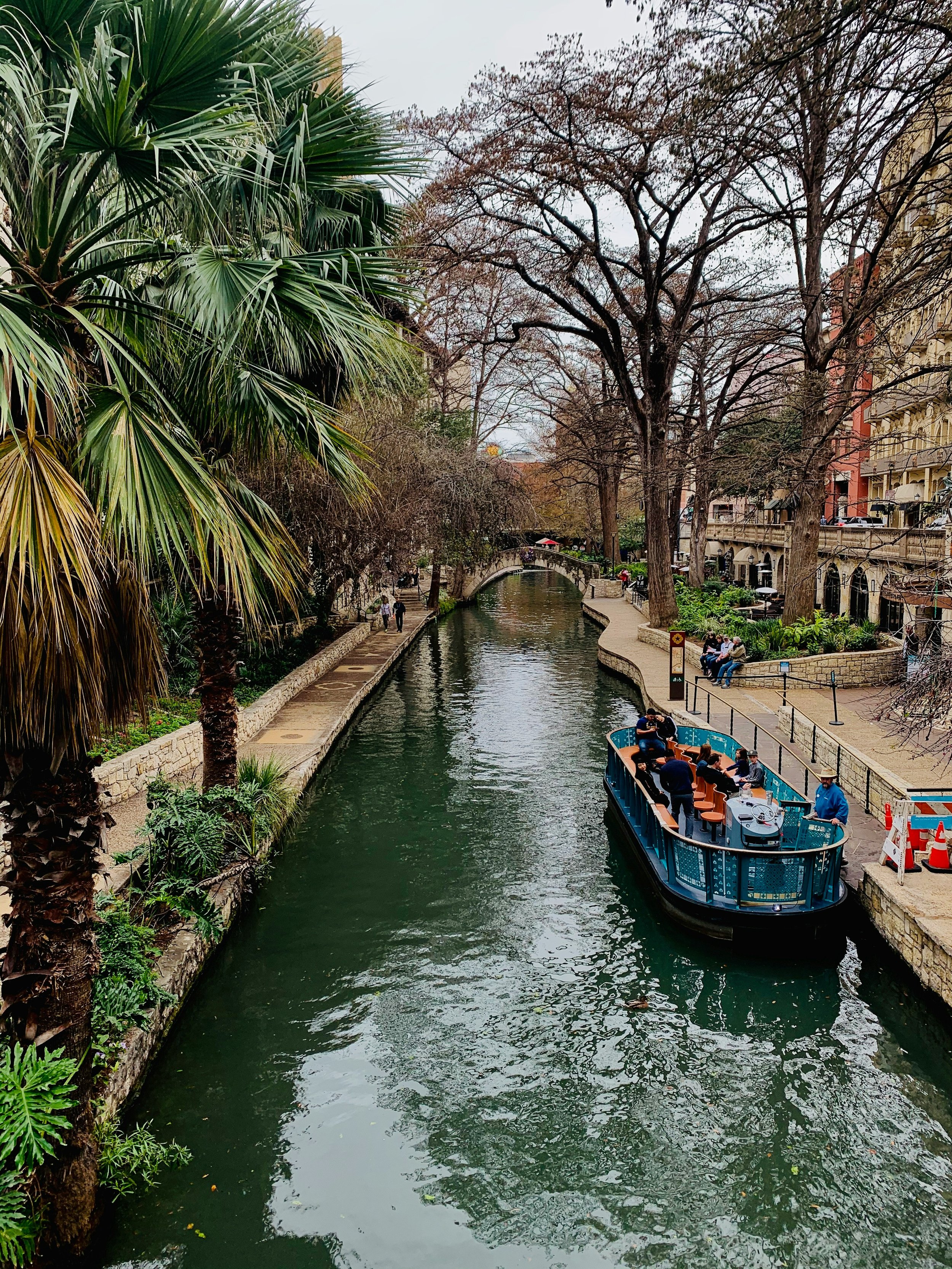 A boat sits on the San Antonio river, where its passengers are served drinks. The river stretches out into the distance with bridges crossing it. The River Walk is lined with trees