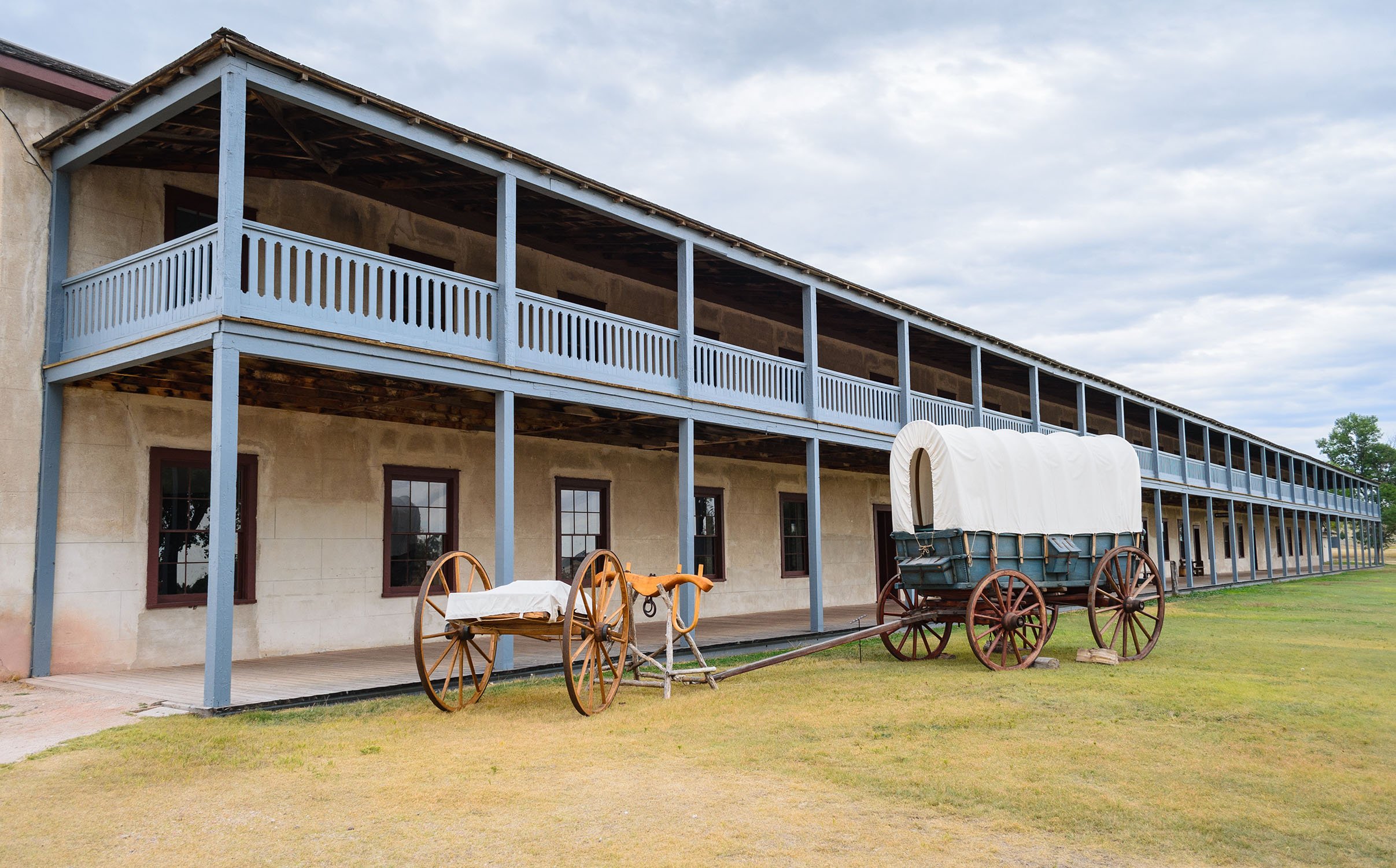 Old fashioned carts and wagons outside a stone building with a long balcony in Wyoming. Drives & Detours Fort Laramie tour