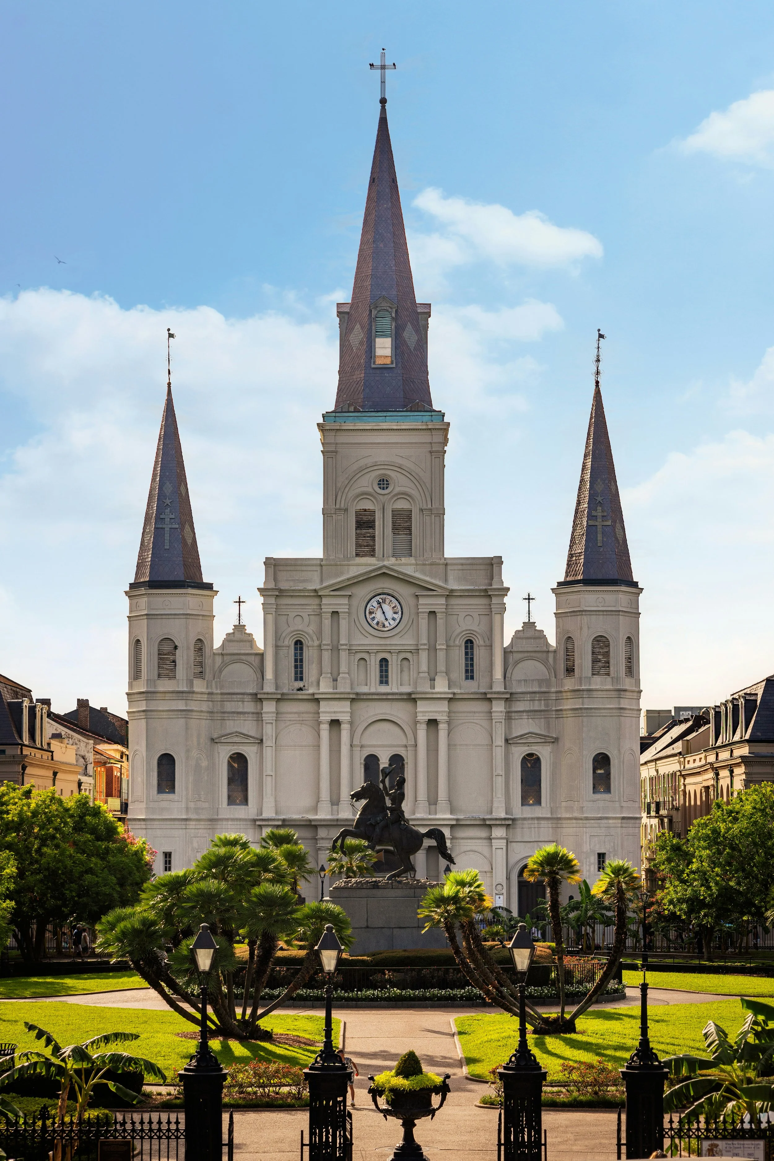 the white St. Louis Cathedral in New Orleans seens from Jackson Square. The cathedral has three spires, and the park is full of green trees and shrubs. A statue of Andrew Jackson on a horse stands in front of it