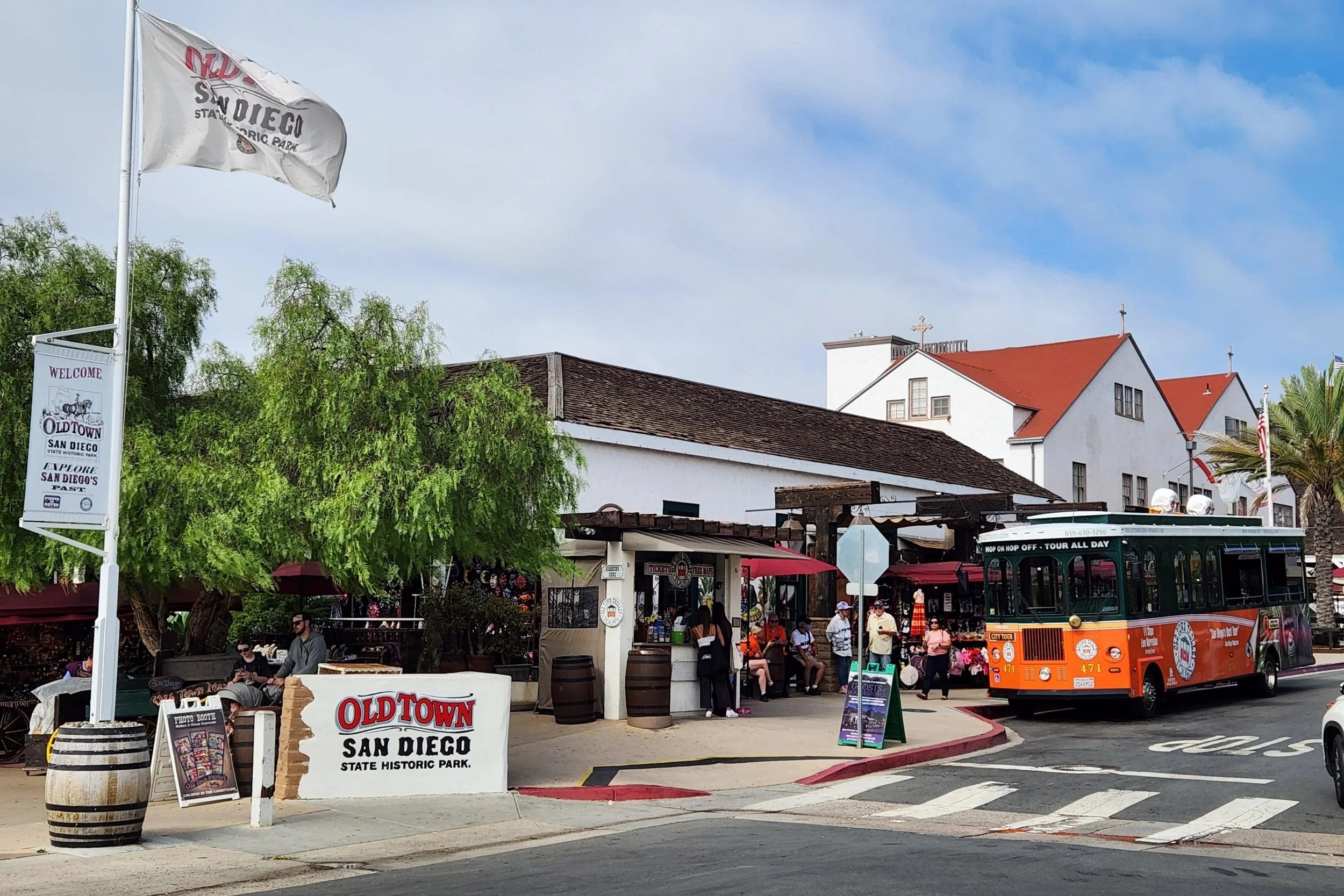 A flag flies over shops and cafes in Old Town San Diego Historic State Park. People mill around and a trollet bus is parked waiting for a tour