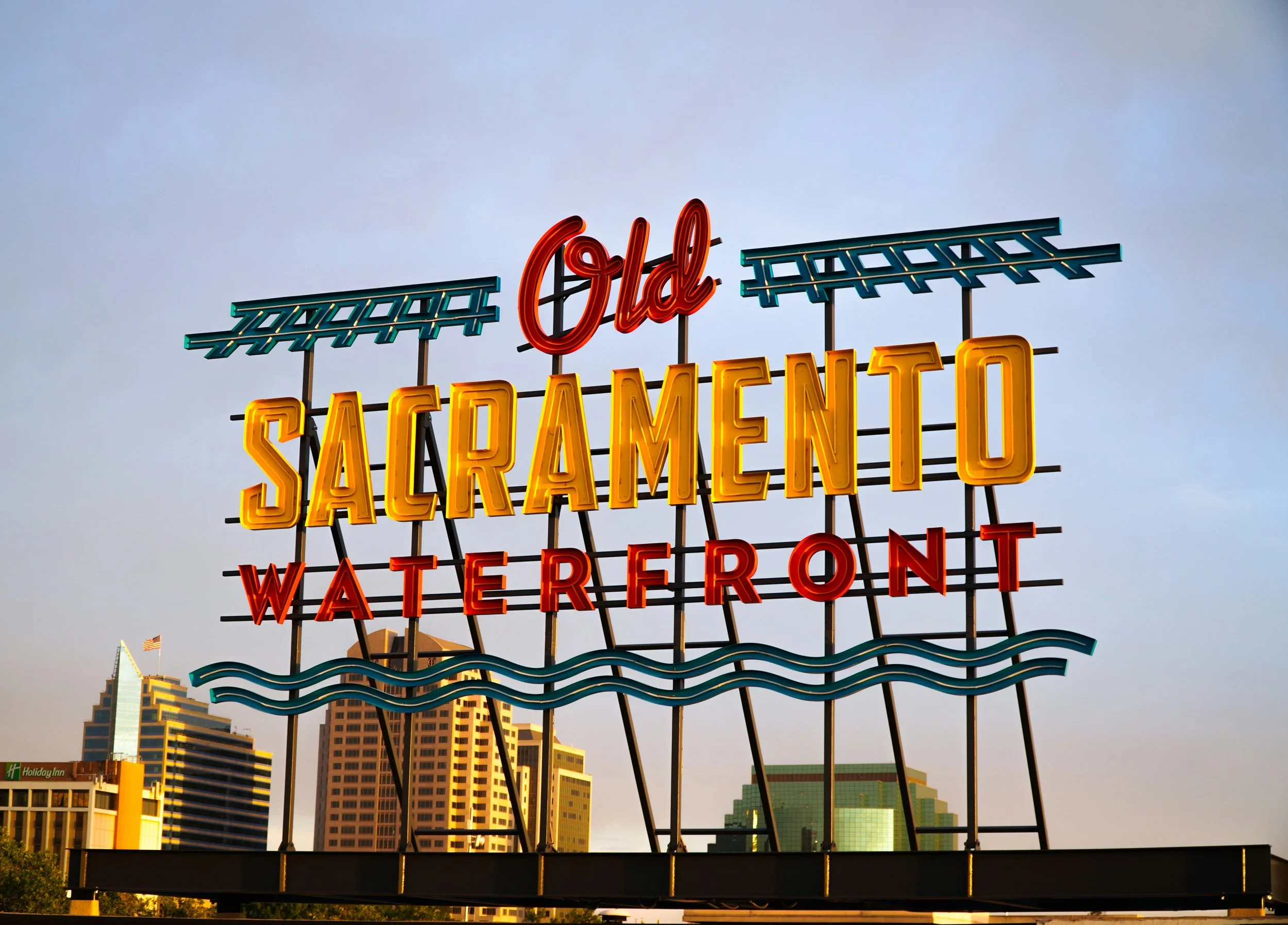 The neon sign of Old Sacramento Waterfront, with train tracks and waves surrounding the text. There are some skyscrapers behind it in the distance