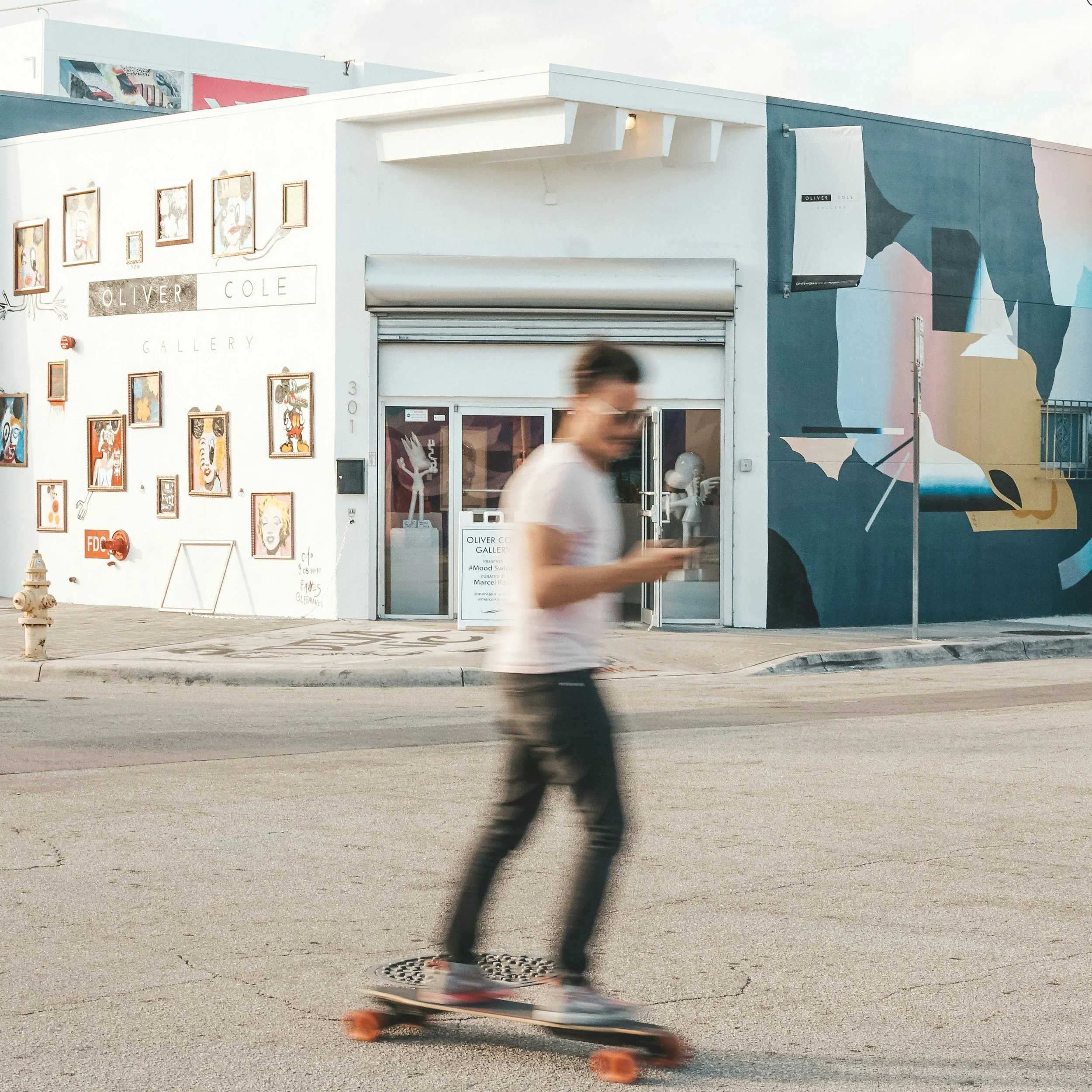 A man skates through Wynwood, Miami. He's out of focus. Street art covers the wall of the Oliver Cole Gallery behind him