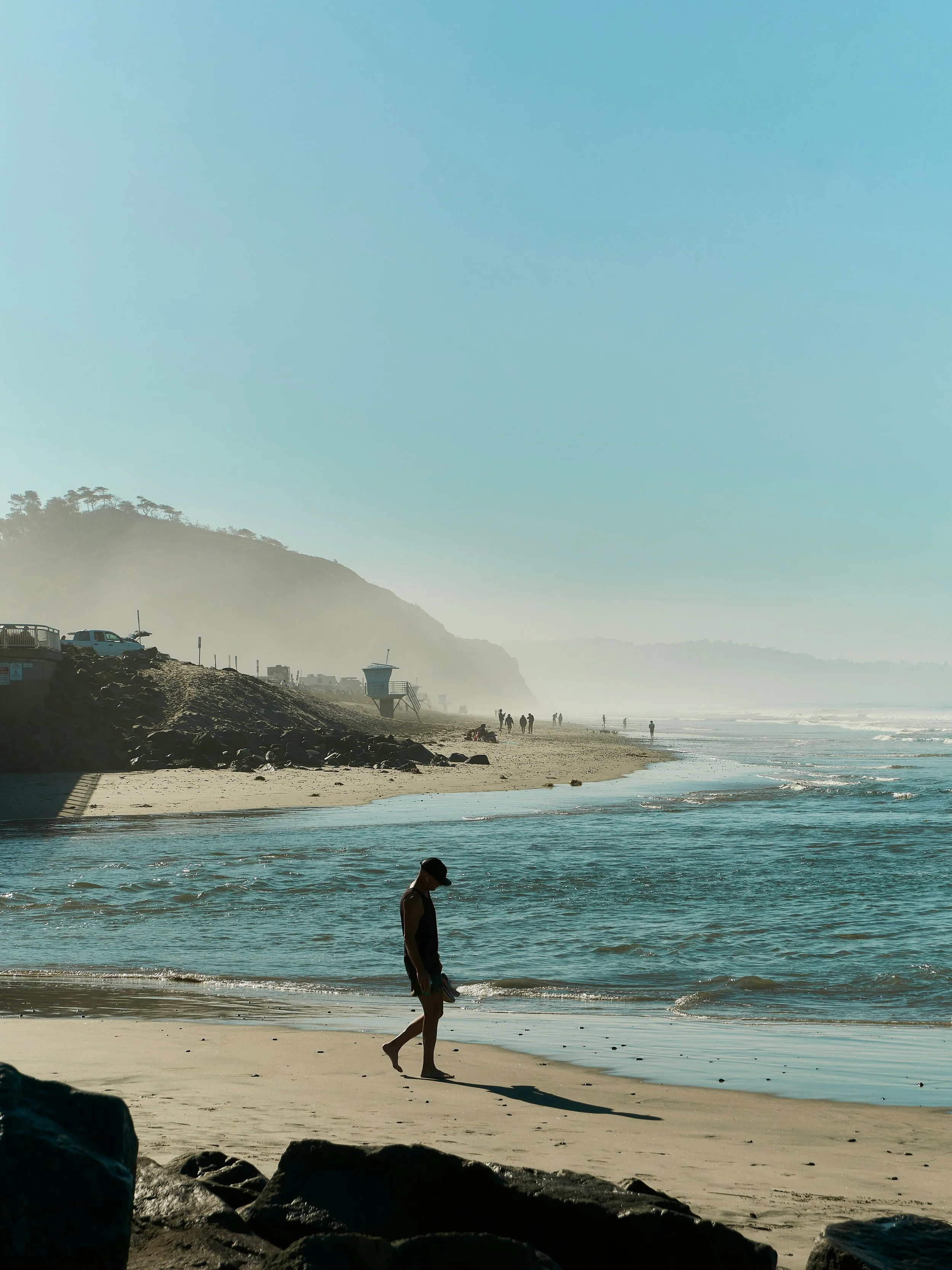 A man walks along Torry Pines State Beach. The sea is lapping against the shore. In the distance, there's a lifeguard hut and cliffs rising out of the sea spray