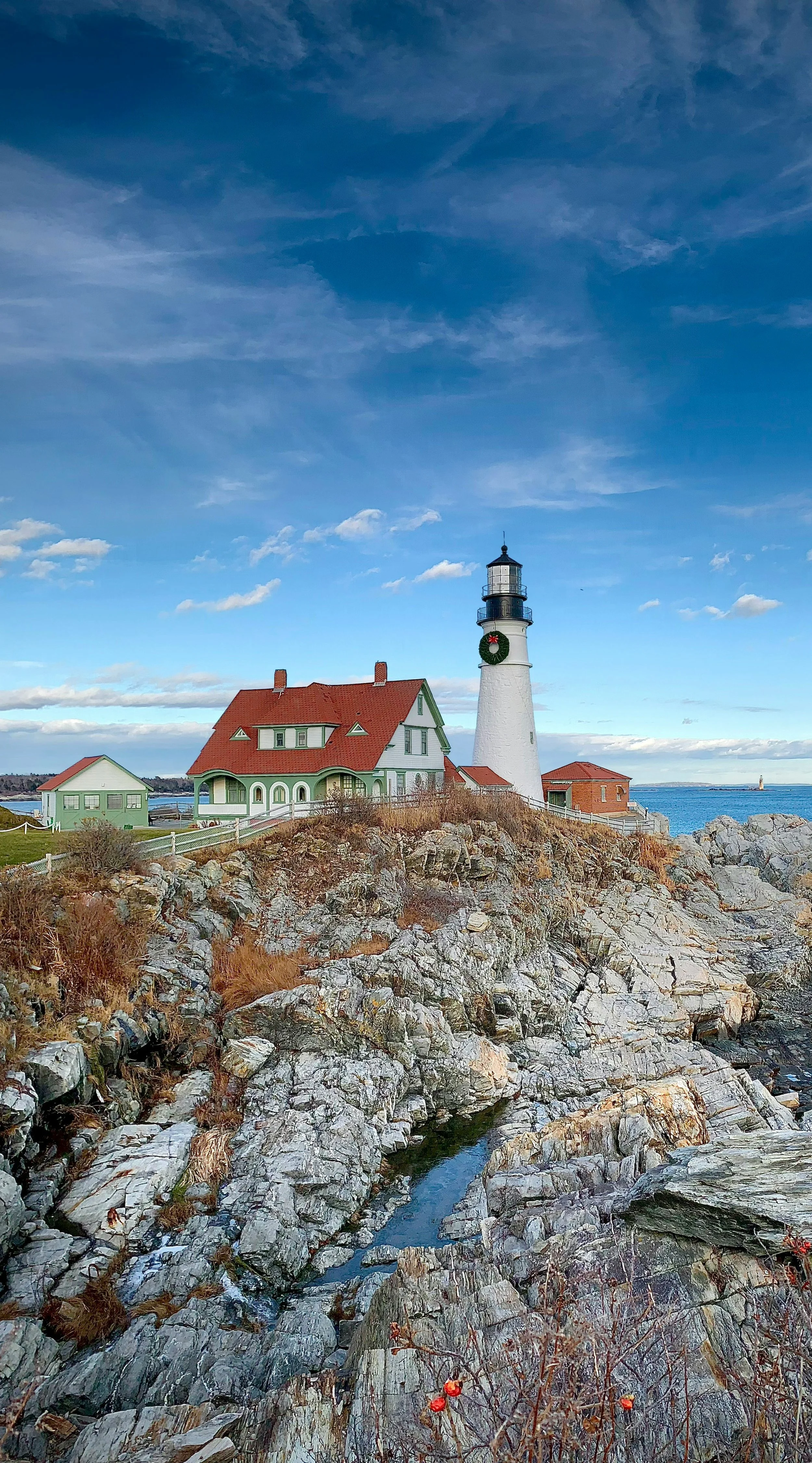 A large white lighthouse can be seen built on some rocks in Portland, Maine. Beside it is a green and white house with a red roof