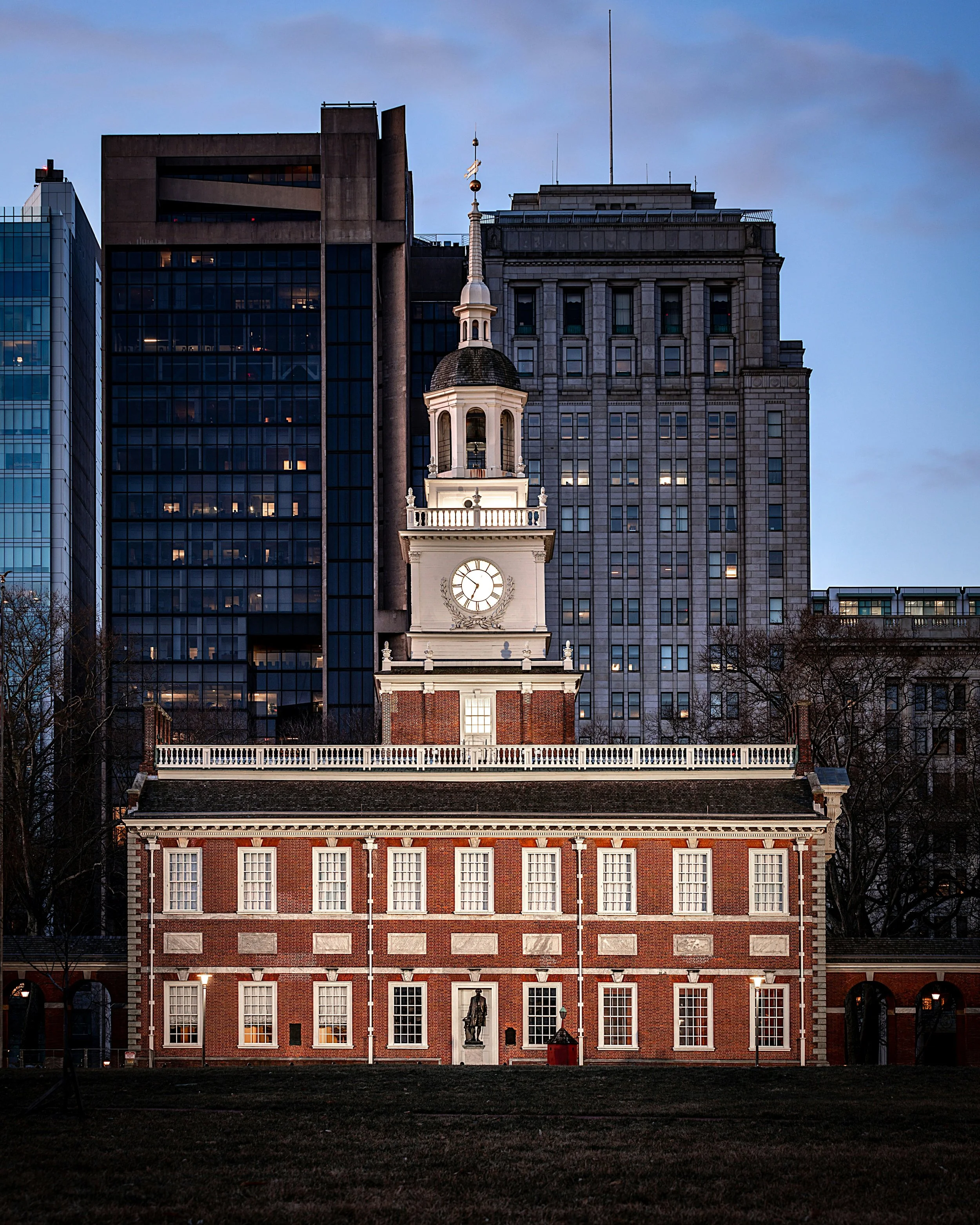Independence Hall in Philadelphia is lit up at night. The red brick building with a white clock tower stand out against the dim skyscrapers behind it