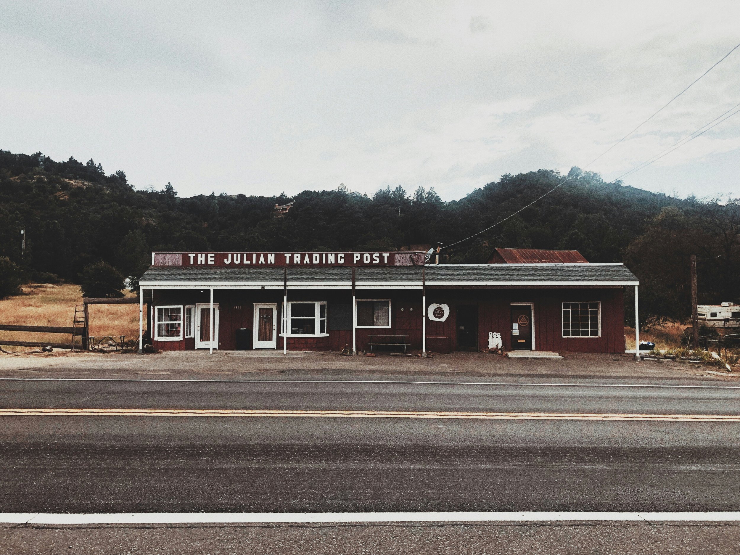 A picture of a single-storey wooden building surrounded by forest. A sign on the roof reads: "The Julian Trading Post." The building is painted brown with white lettering and details
