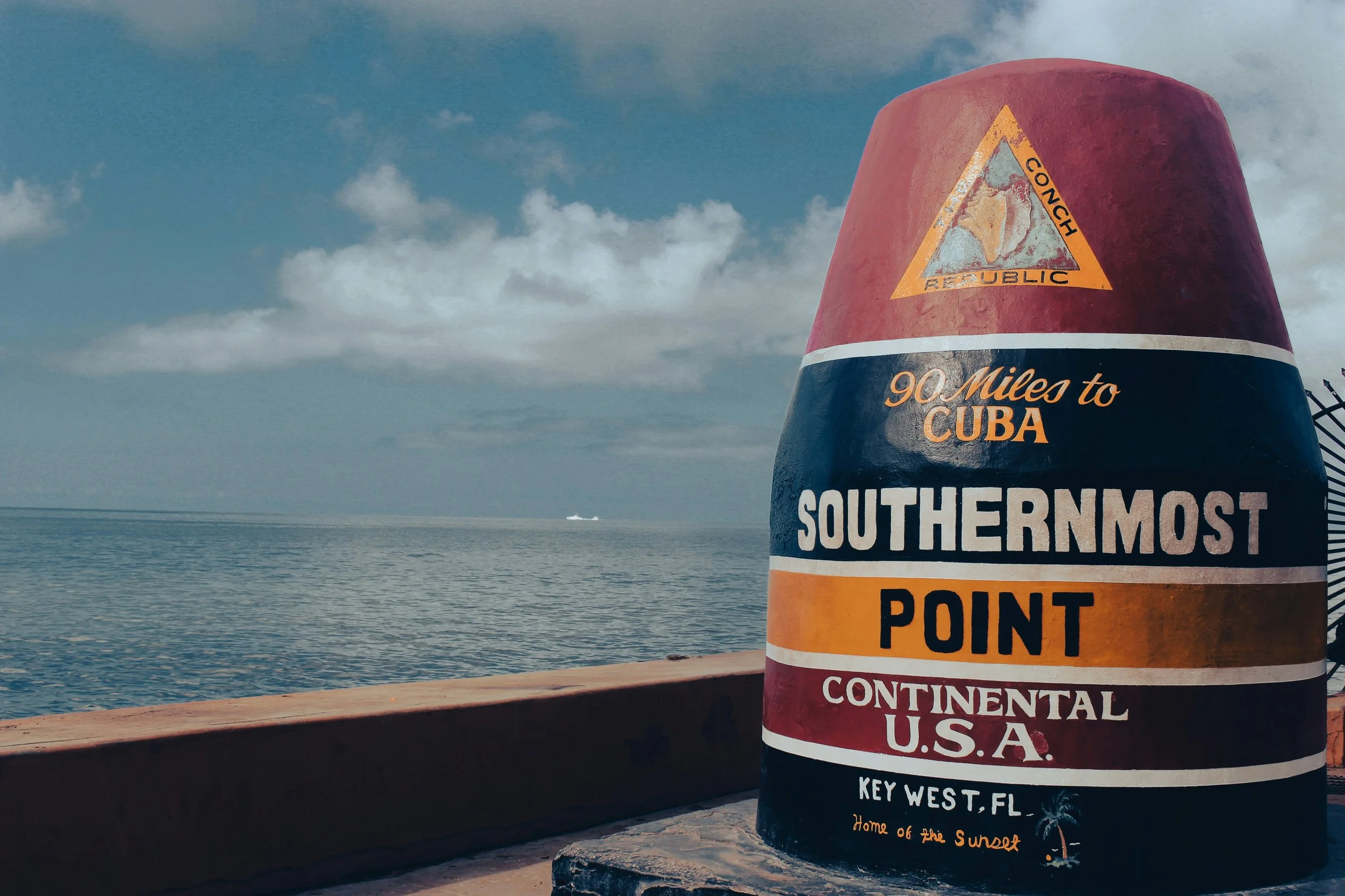 A buoy installed on land at the Southernmost Point of the Continental USA in Key West, Florida. It is painted in dark blues, reds and yellows. The ocean stretches out behind it