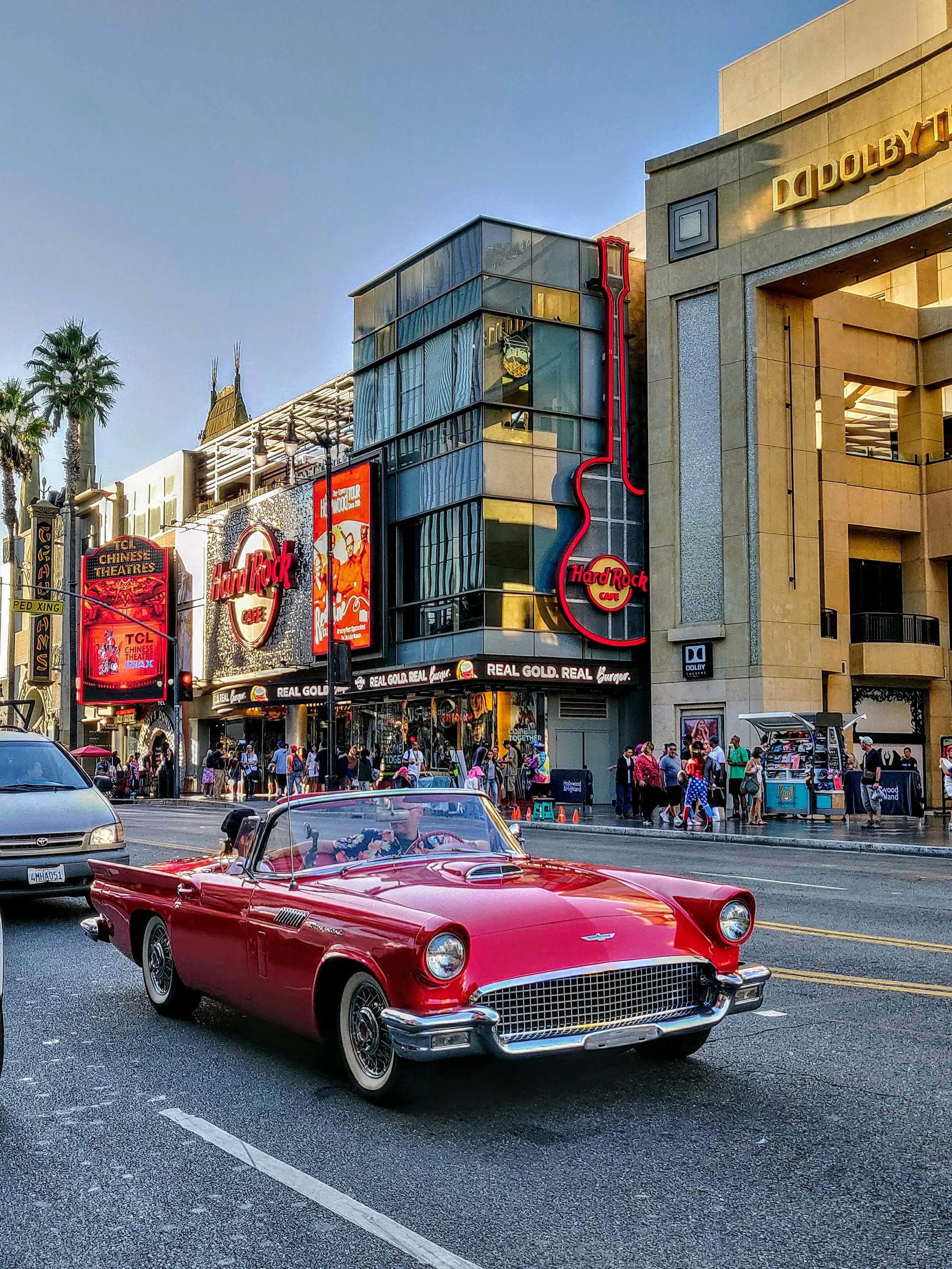 A red, classic Chevrolet Corvette drives past the neon signs of Hollywood Boulevard