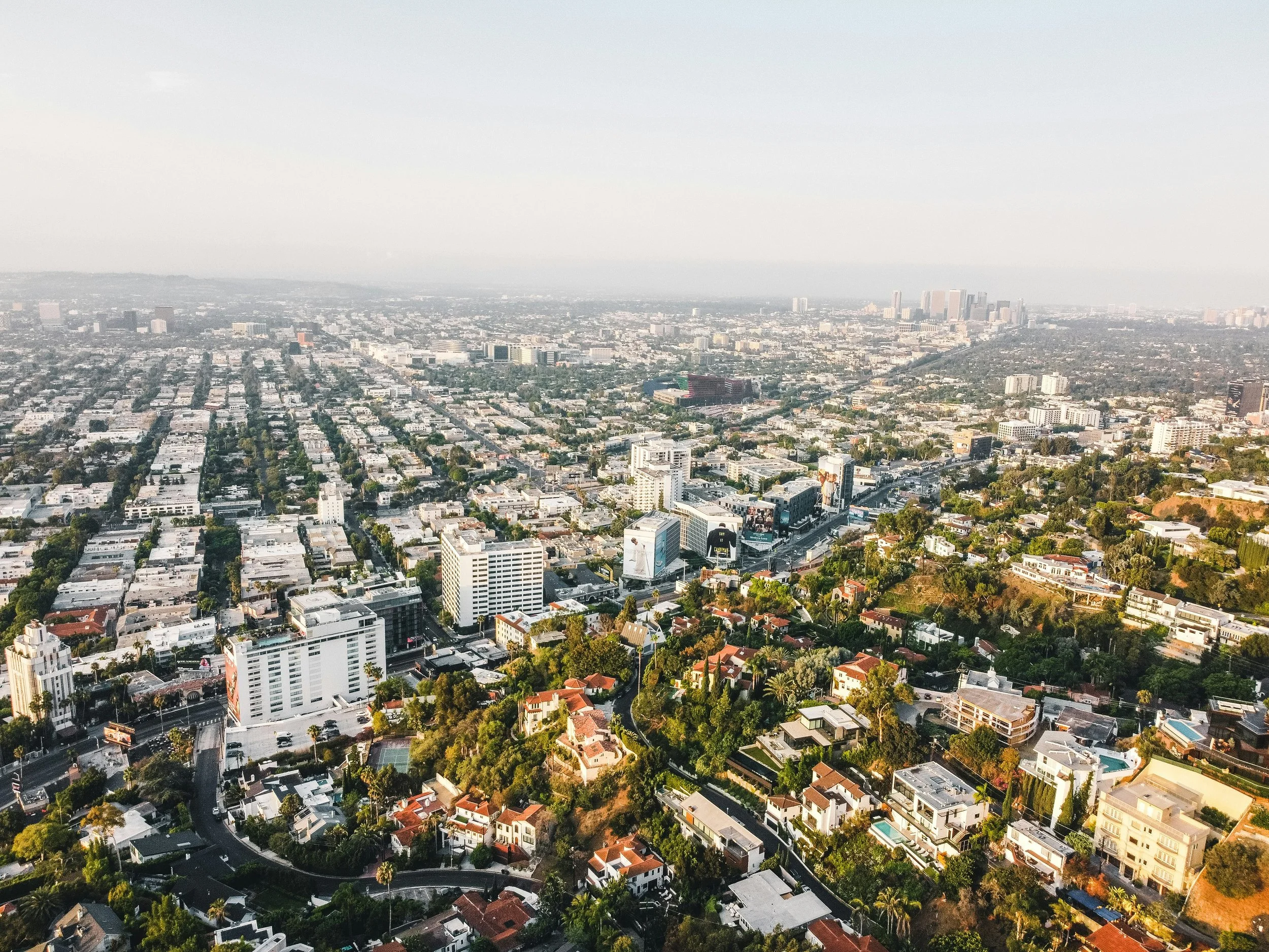 An aerial view of Beverly Hills, Los Angeles. The city looks mostly flat, apart from the greener area that climbs towards the camera. Downtown LA is visible in the distance