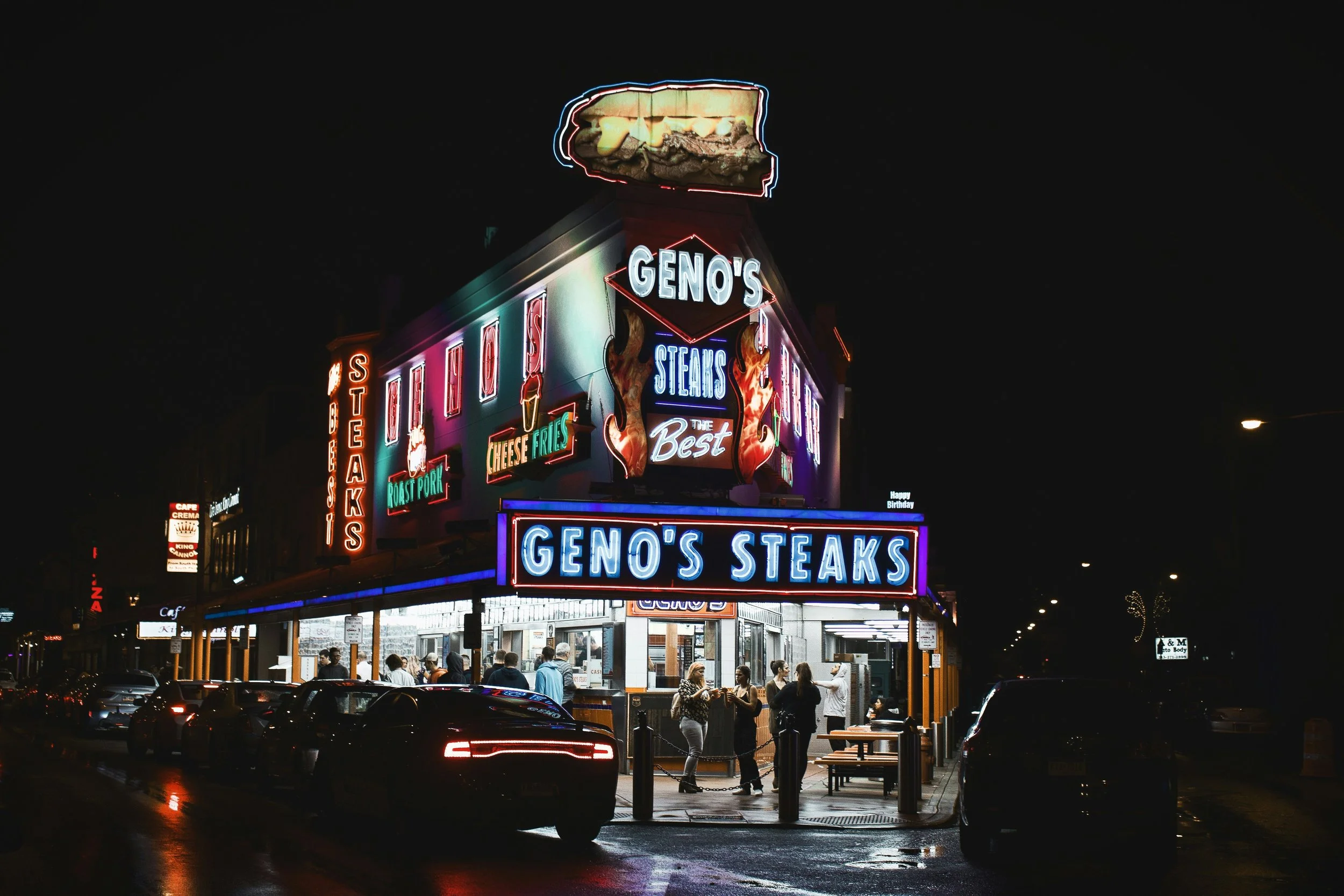 A restaurant is covered in neon signs at night. They read: Geno’s Steaks. Drives & Detours first time in Philadelphia