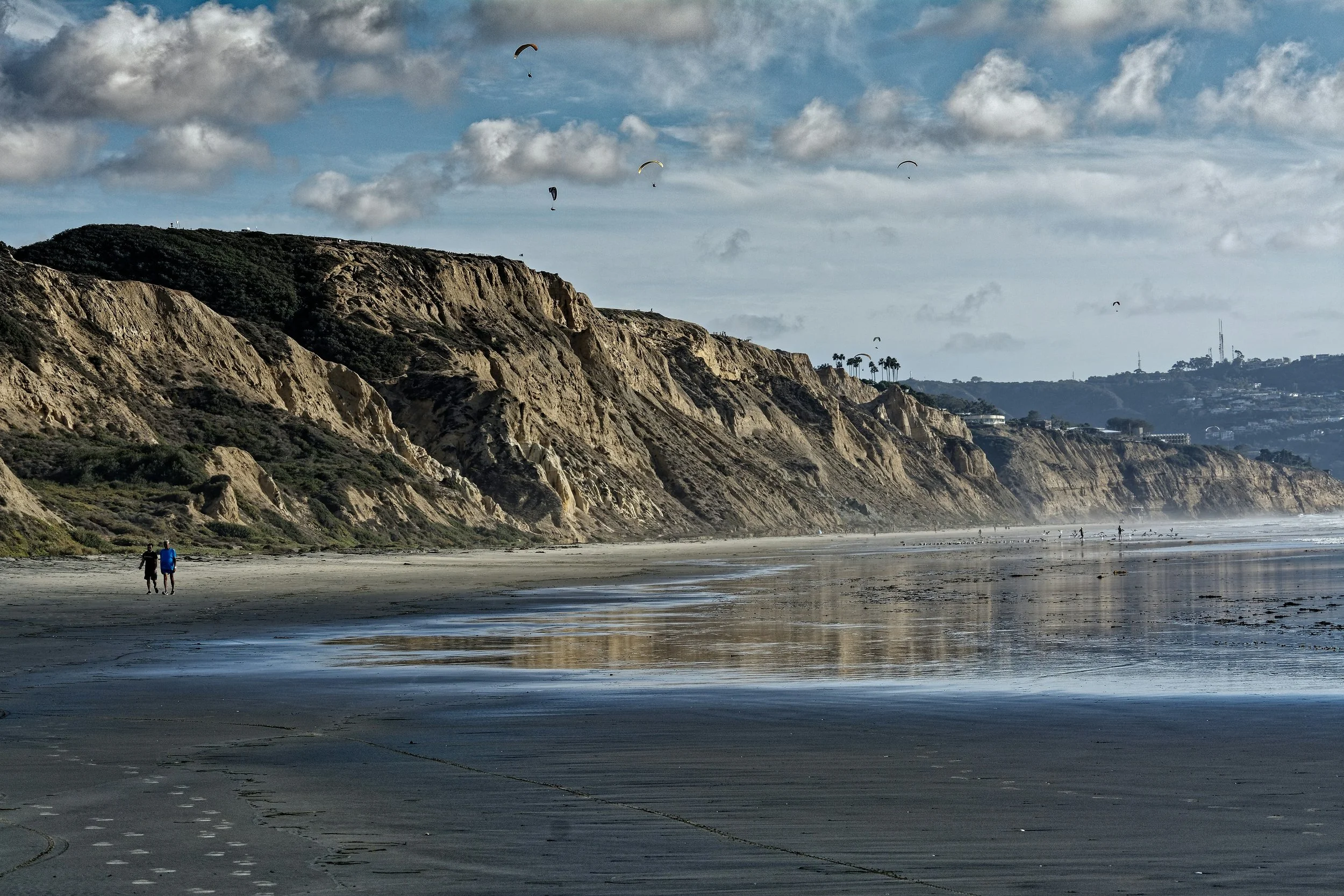 Hang gliders float over cliffs that run down to the sea. Two people walk along the waters edge, as if they are enjoying their San Diego Driving Tour