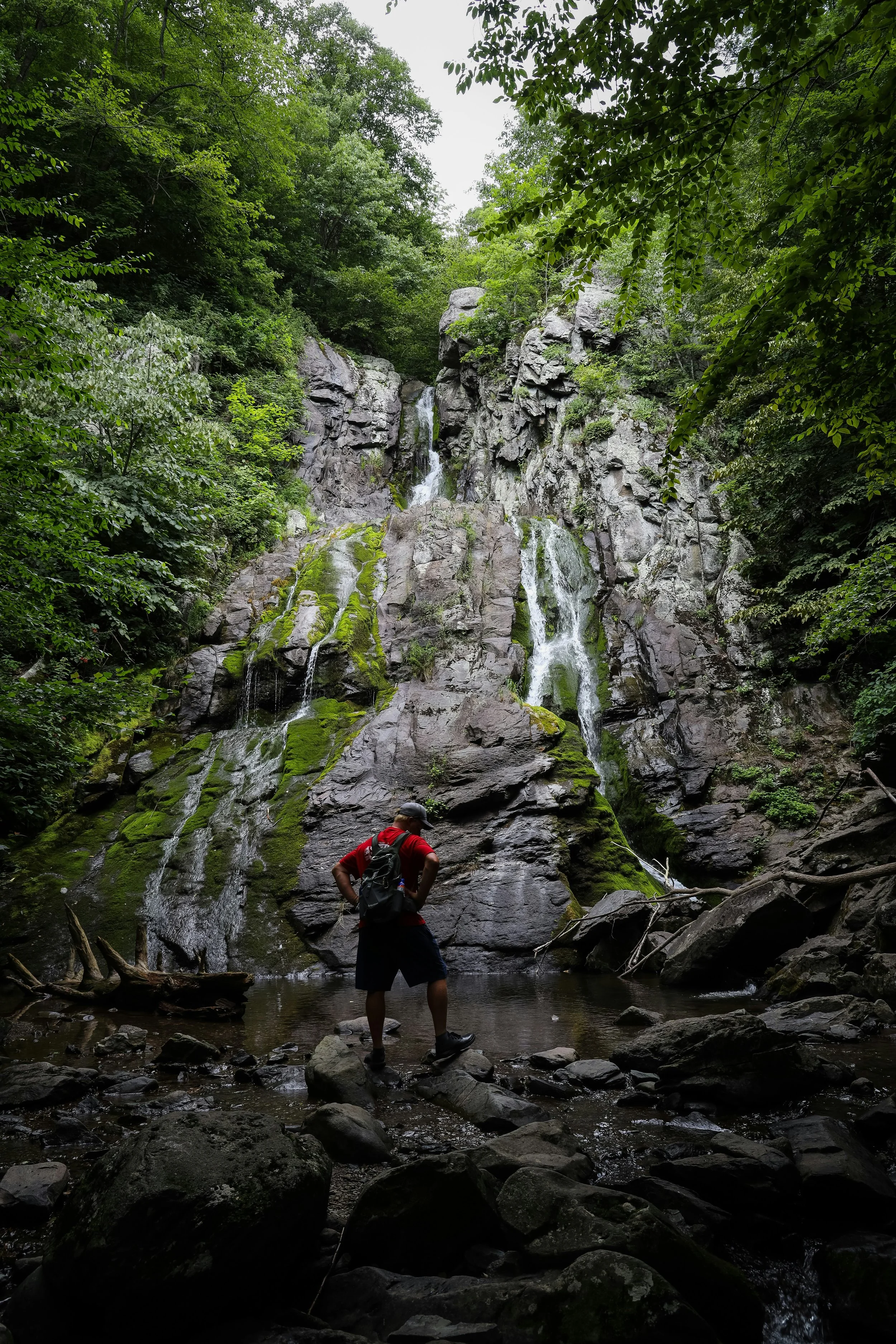 A man in a red t-shirt, carrying a rucksack, stands at the bottom of a waterfall that tumbles over a rockface into a pool in Shenandoagh National Park, near Skyline Drive