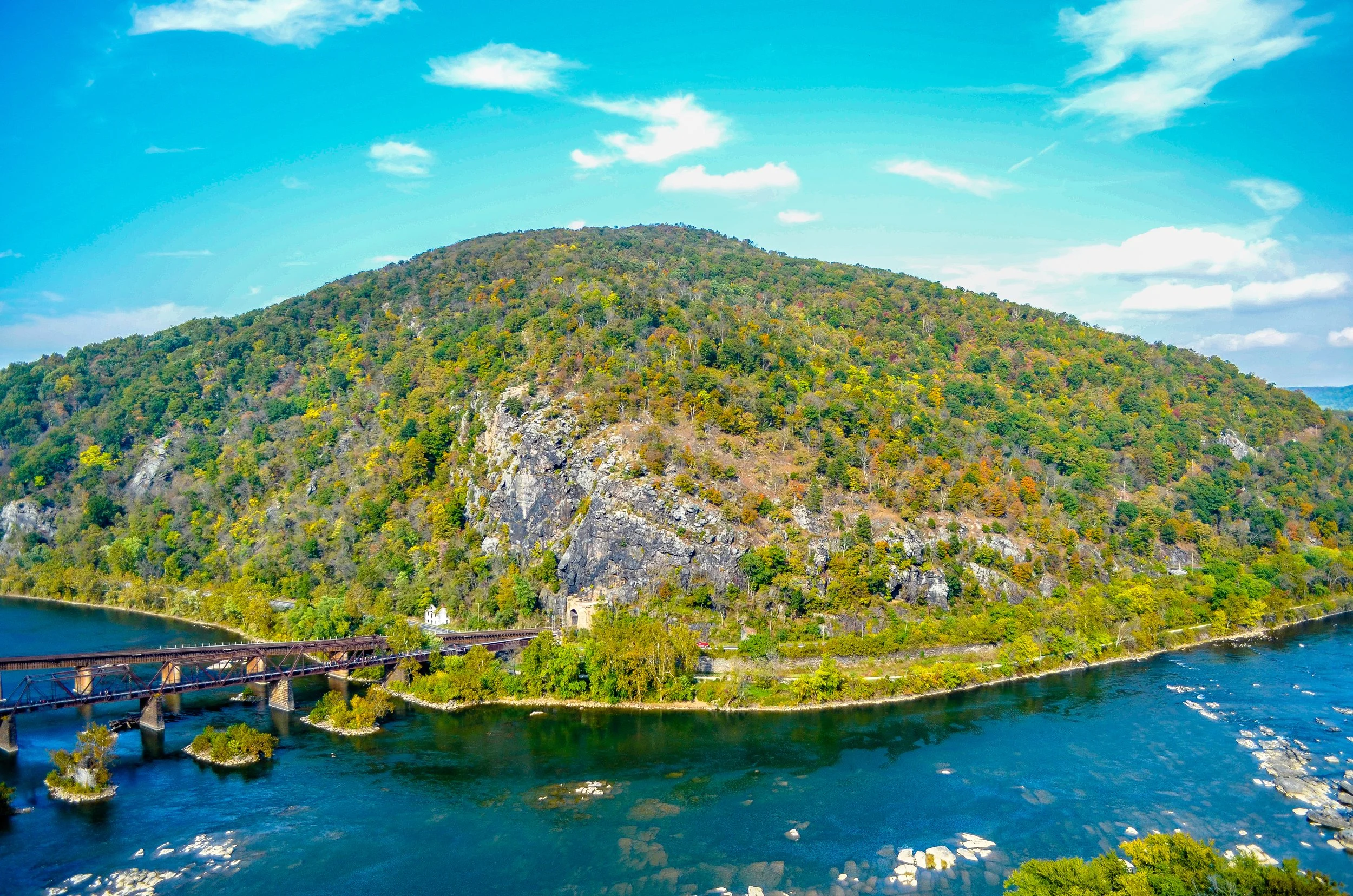A rail line runs across a bridge over the Potomac and Shenandoagh Rivers towards a tunnel in a cliff. Drives & Detours Harpers Ferry hidden gems