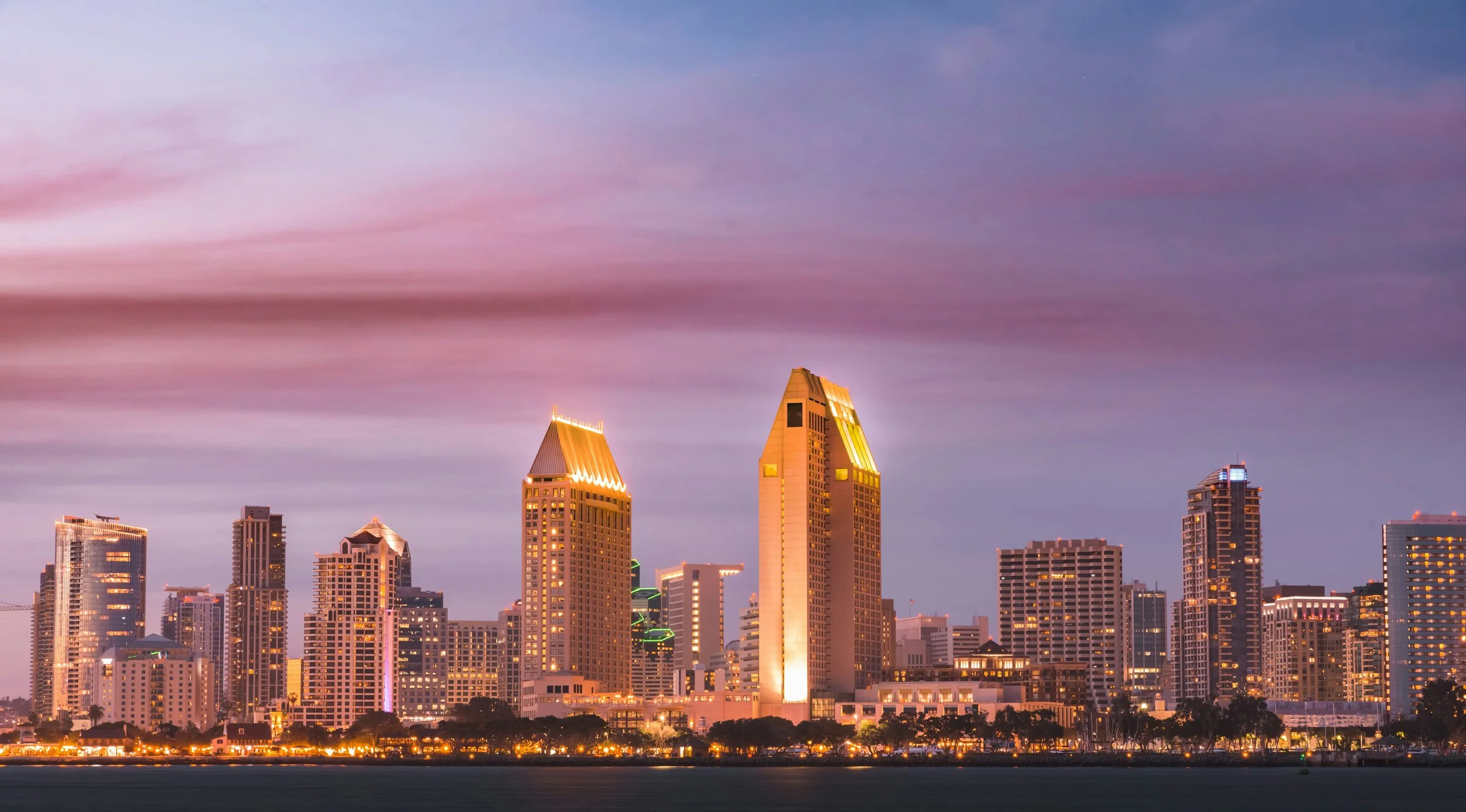 The San Diego skyline is lit up at sunset. The skyscrapers are bathed in soft light from the sky which is a gradient from orange to blue
