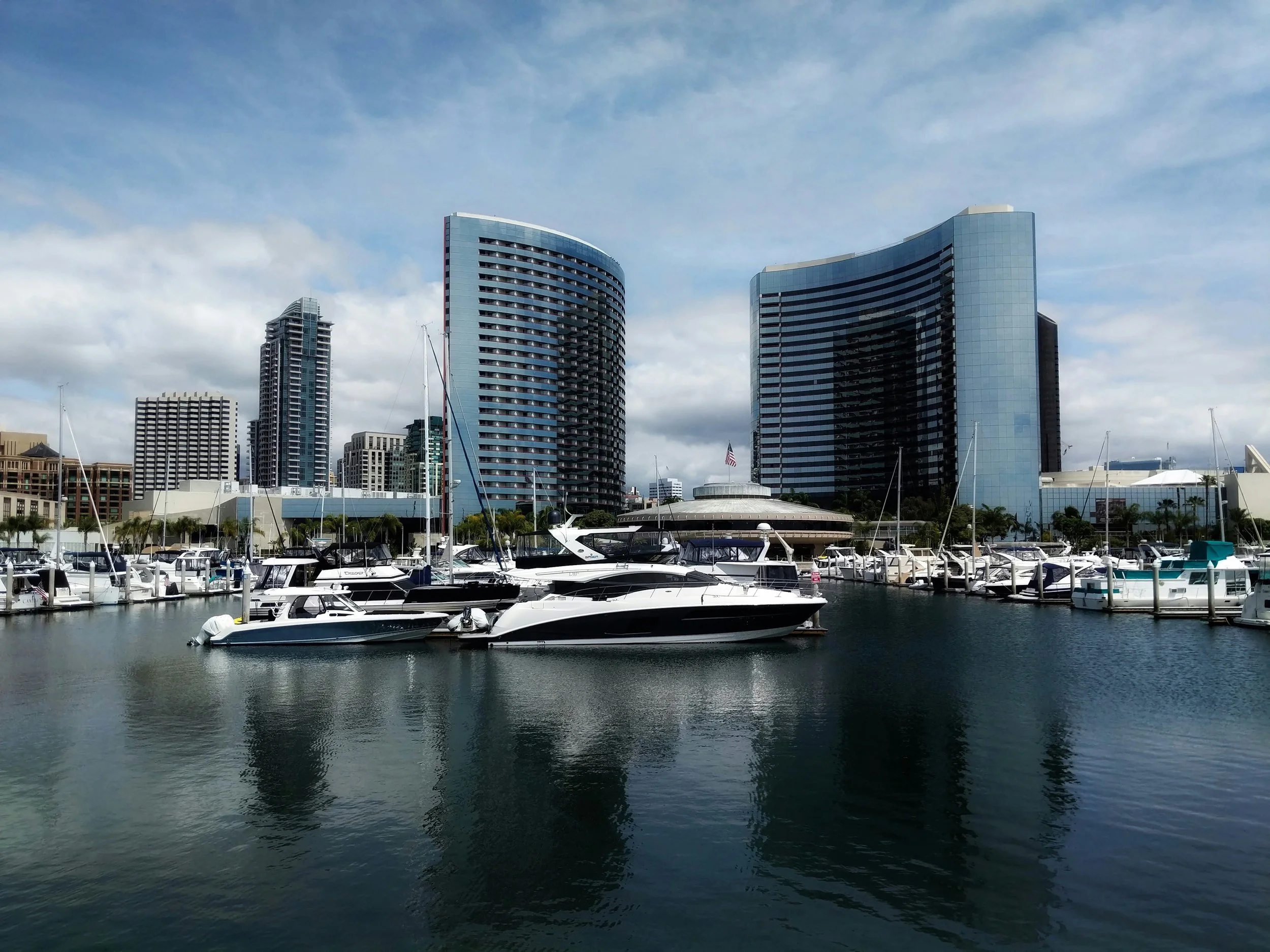 Modern yachts float on still water in San Diego Bay under skyscrapers. Drives & Detours USS Midway Museum and Embarcadero Self-Guided Tour