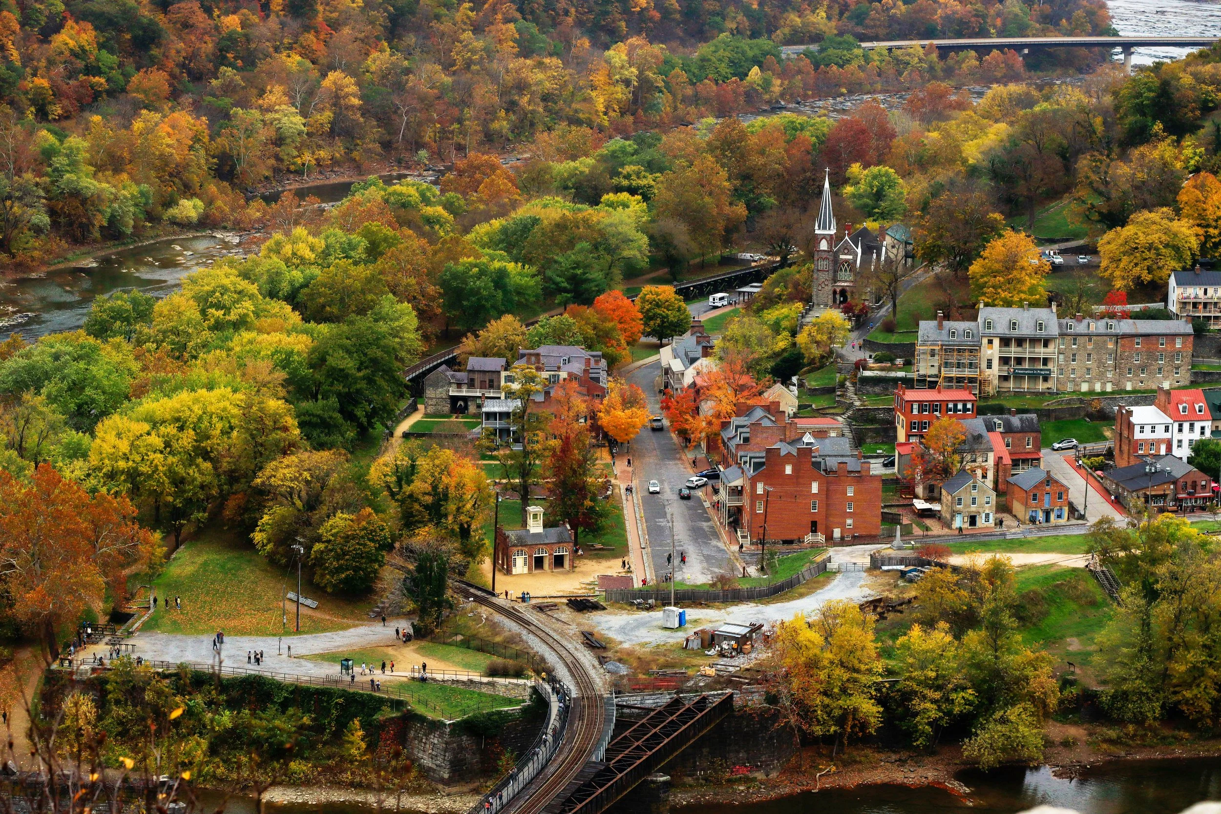An aerial view of Harpers Ferry. It's possible to see the railroad crossing, churches and old buildings. The town is surrounded by the autumnal colours of the forest and rivers