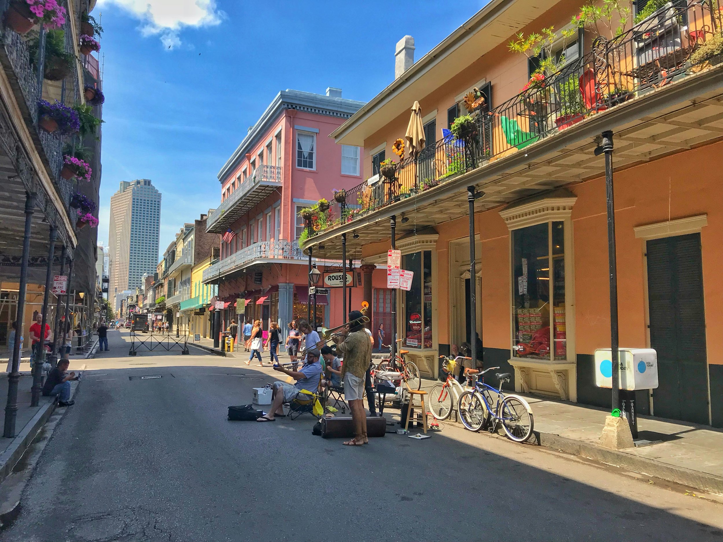 A jazz band plays on the street in New Orleans during daytime. The are surrounded by buildings with iron balconies. A few cruiser bicycles sit beside them