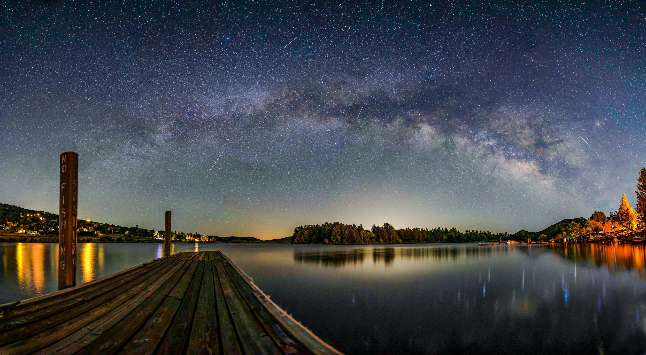 Shooting stars fall through the night sky over a lake in Julian, California. The whole Milky Way can be seen. A copse of trees is reflected in the lake on the far side