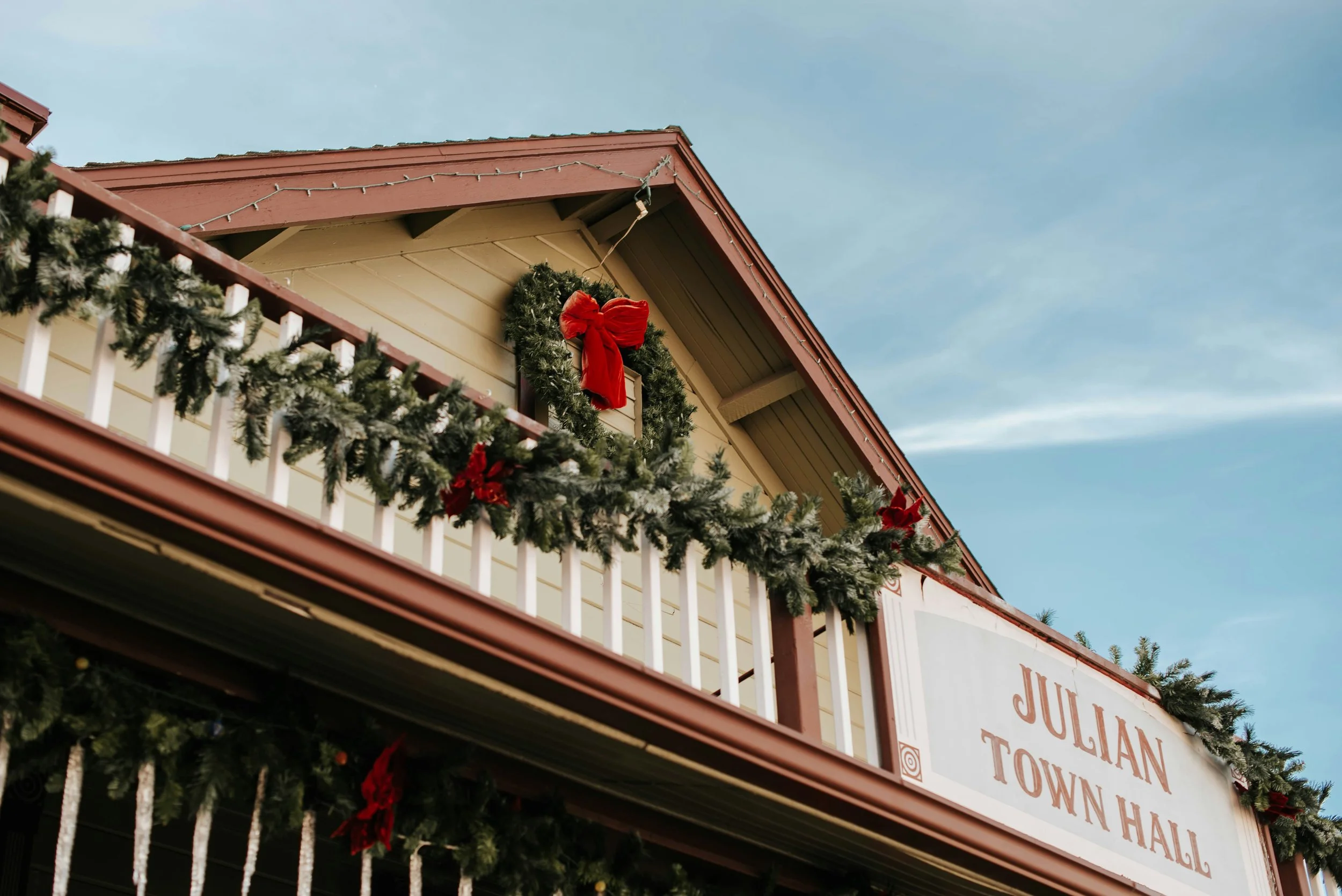 Julian Town Hall is dressed up for Christmas. The picture is a detail of the balcony and roof, which are lined with holly. A holly wreath with a red bow hang from the roof.