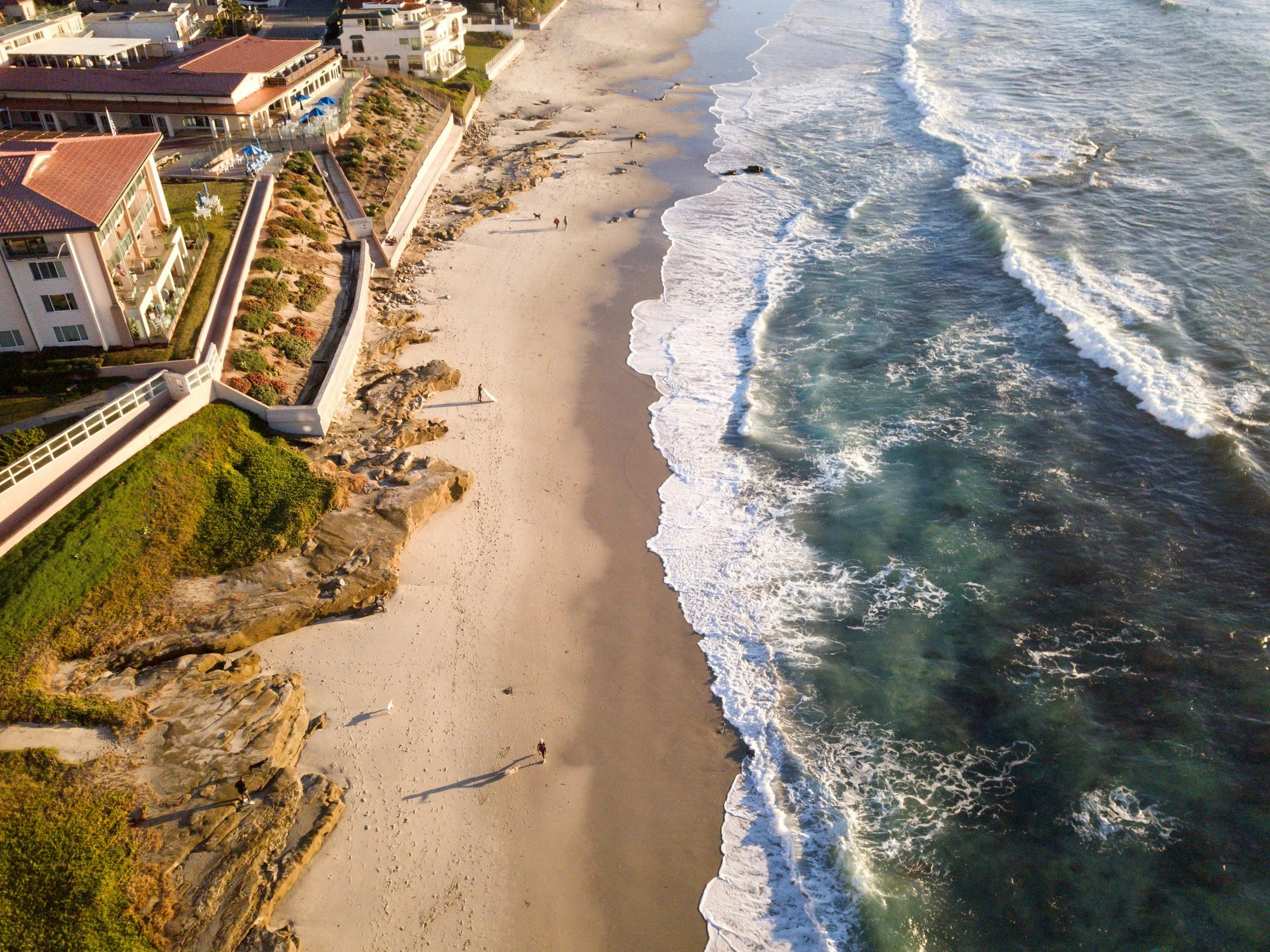 An aerial view of the beach at La Jolla, San Diego. People walk along the sand as waves break against it. Large houses line the edge