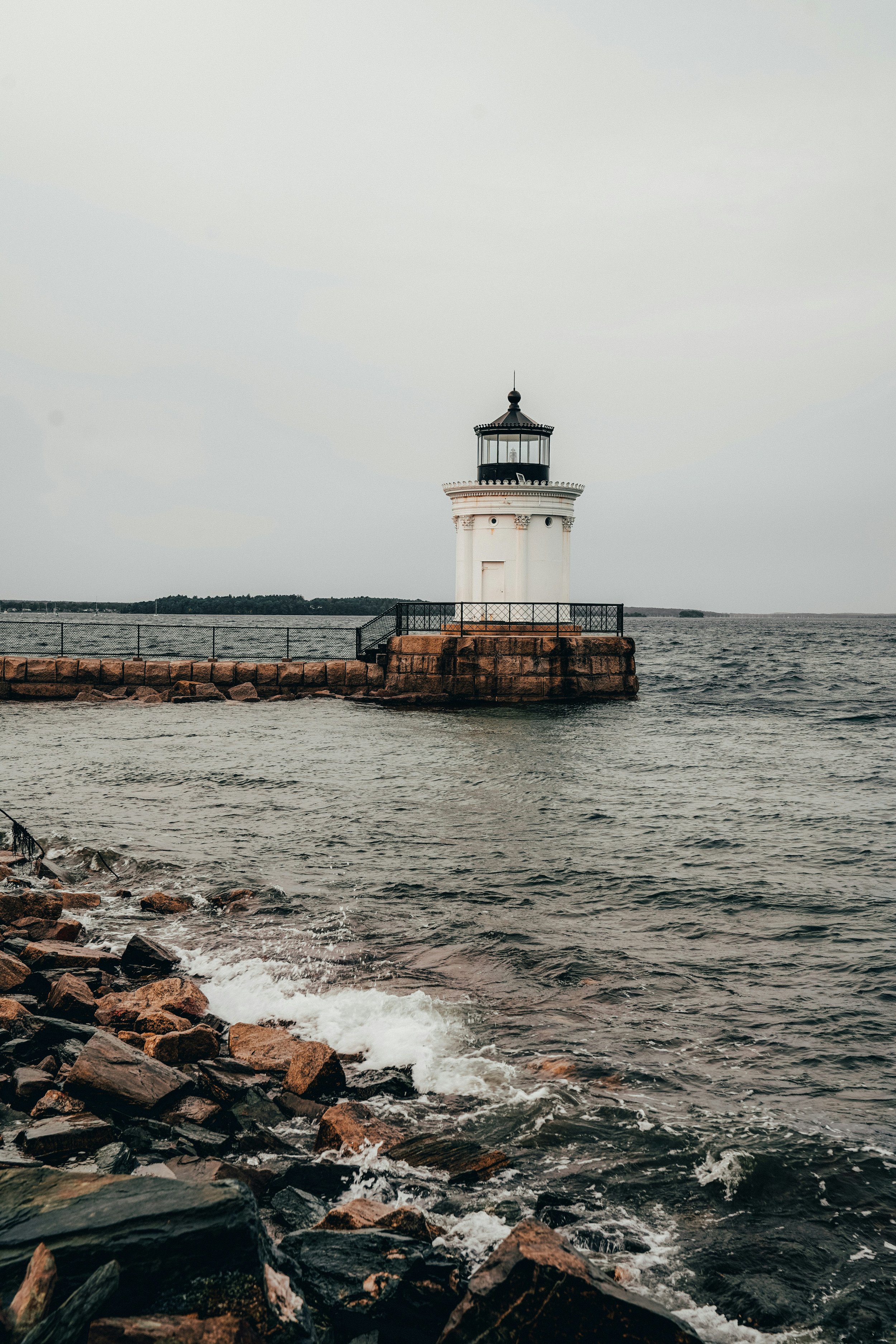 A small, white lighthouse sits in the water in Portland, Maine. Its a cloudy, grey day