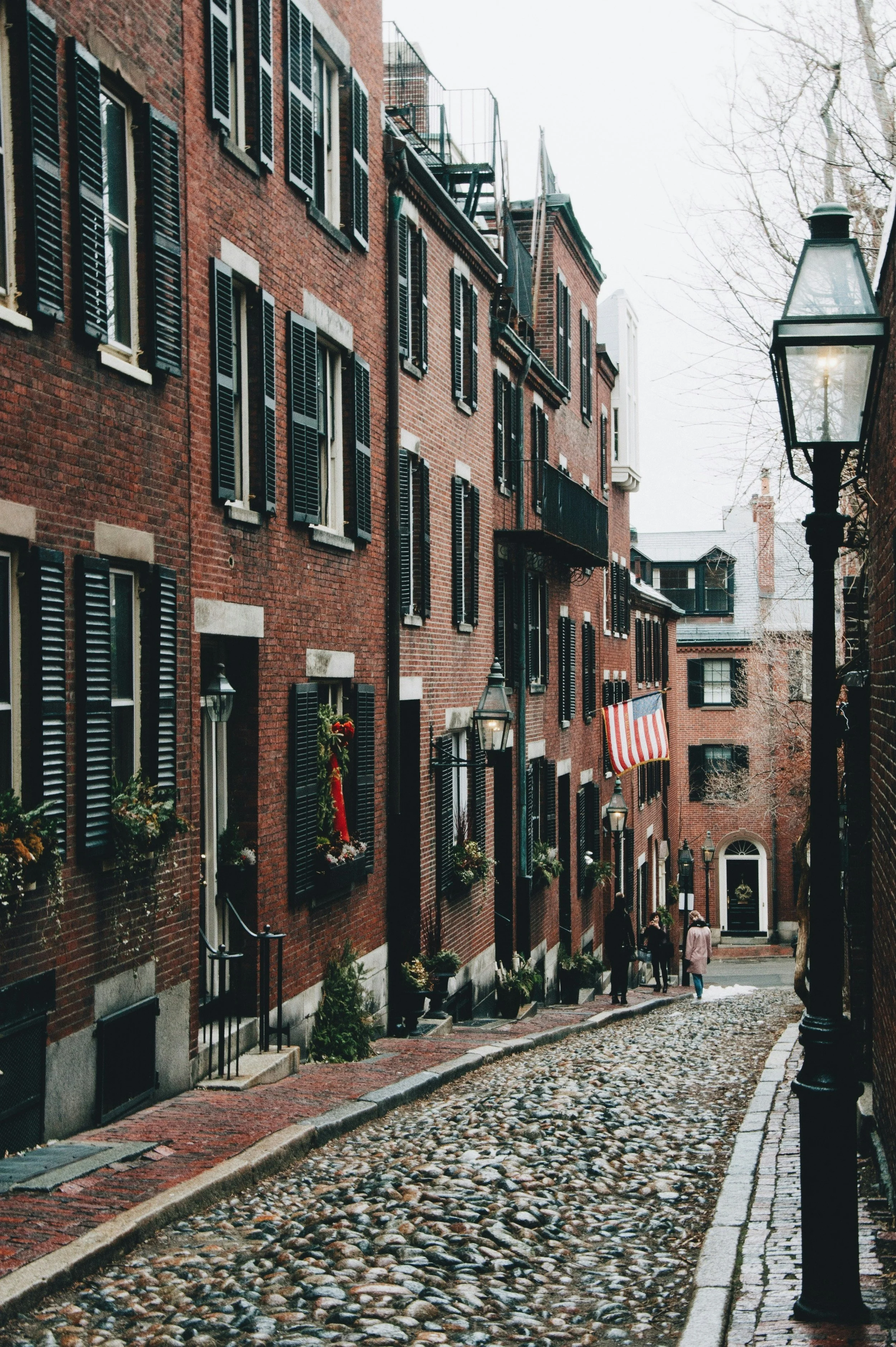 An American flag flies in front of a building on the cobbled Acorn Street in Boston. People pose for pictures in front of the brick terrace houses