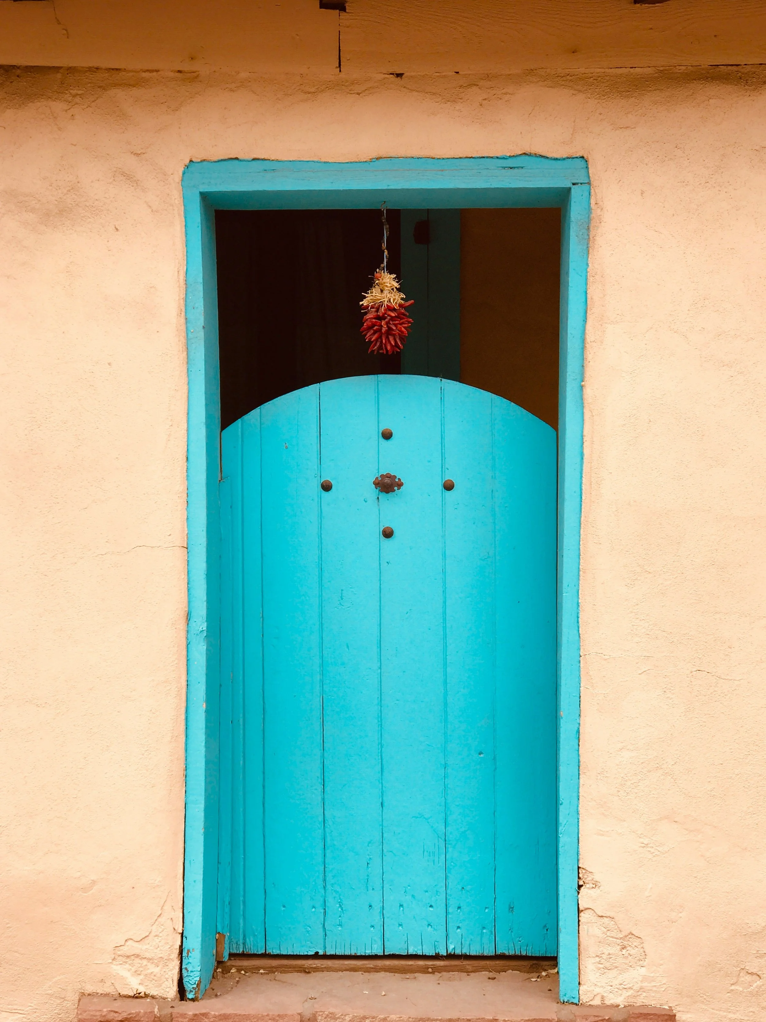 A close up of bright blue door on an adobe building in Santa Fe. Red flowers hang from the top of the door frame