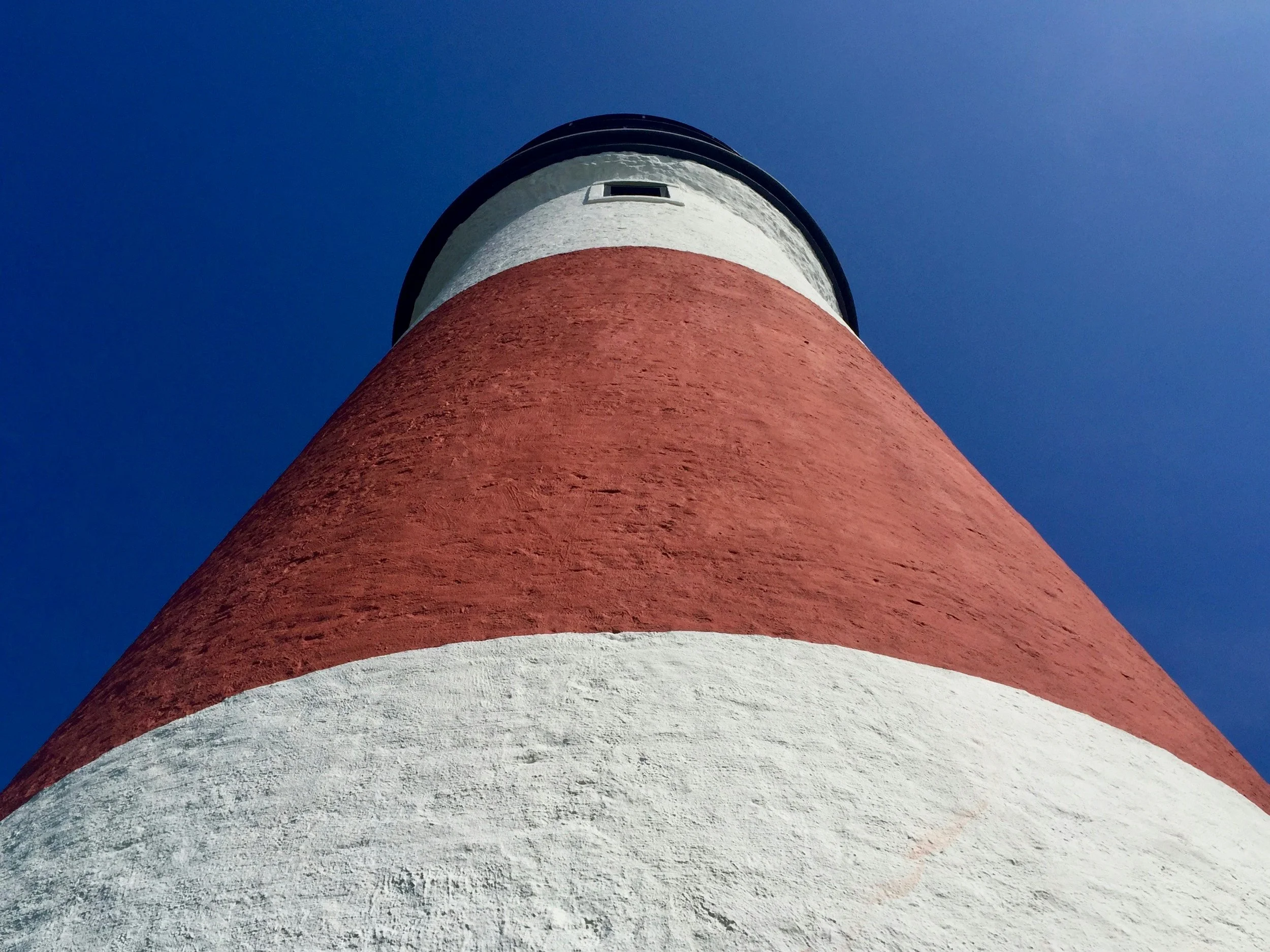Sankaty Head Lighthouse is seen from below at an extrem angle. The thick red and white stripes fill the lower two-thirds of the picture. A small window is at the top, just under the light platform. The sky is a deep blue