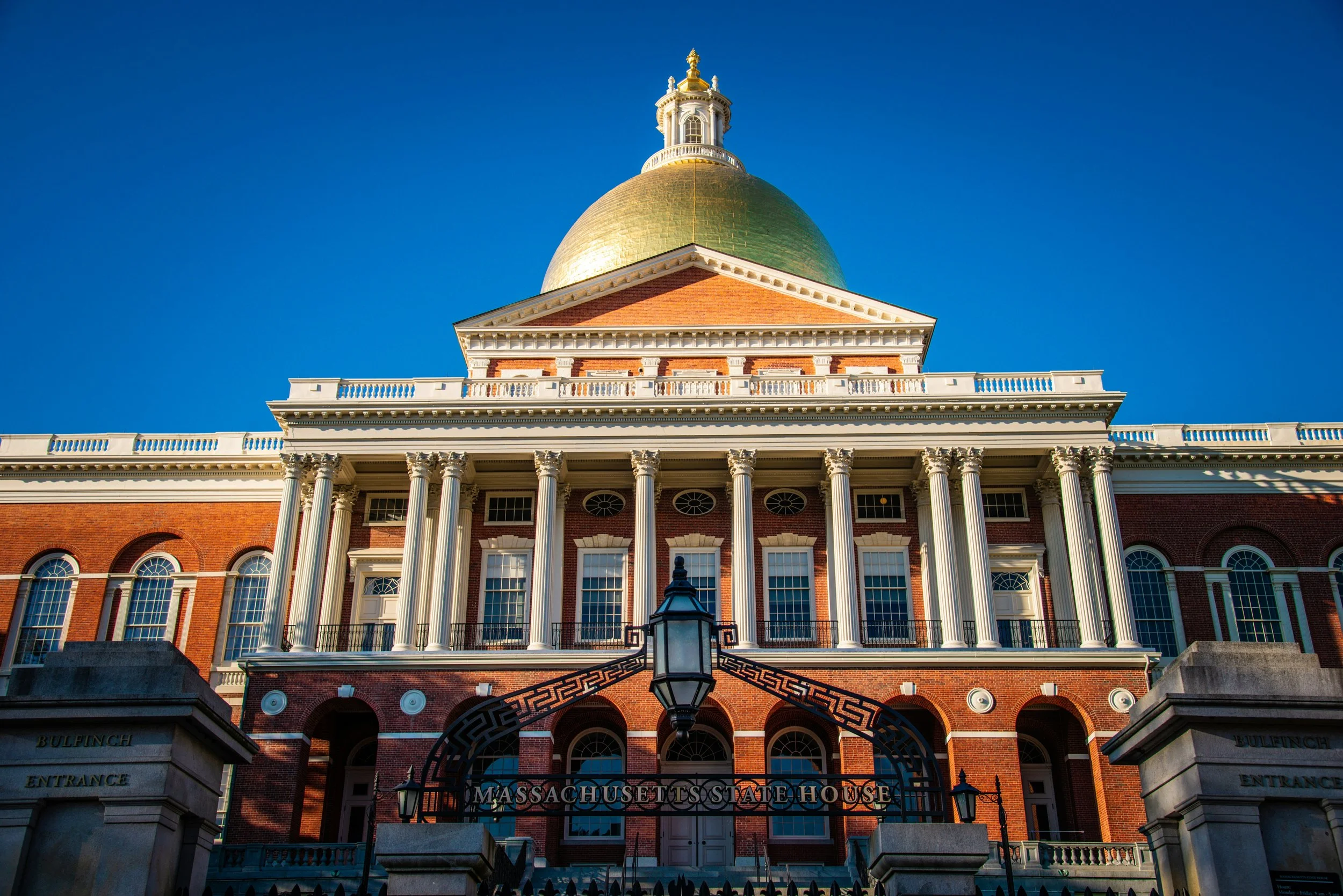 A picture of Massachusetts State house from the front. The sun gleams off its huge golden dome. White pillars decorate the front of the red brick building.