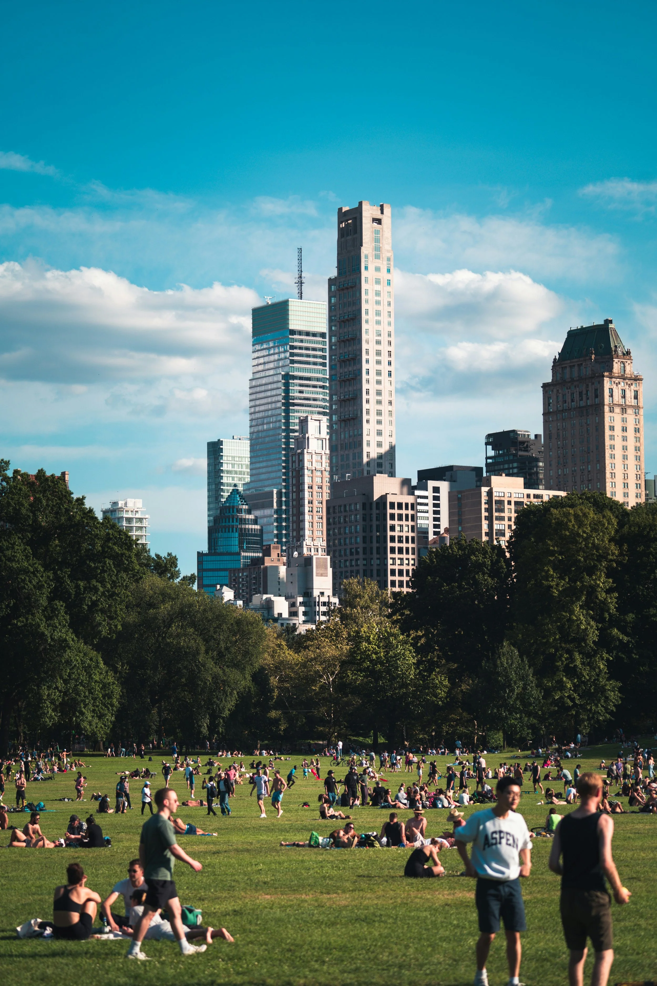 A crowd fills the spaces of Central Park, New York, on a sunny day under the skyscrapers.